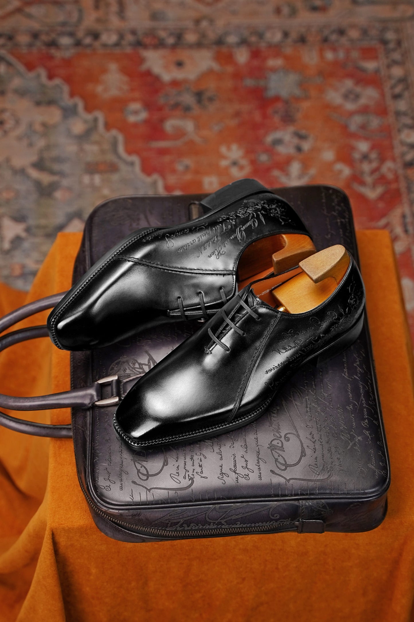 A pair of black leather dress shoes with wooden shoe trees inside, displayed on top of a black embossed leather case over an orange fabric surface with a patterned rug in the background.