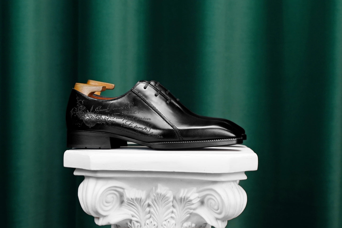 A single black leather dress shoe with embossed floral detailing, displayed on a white decorative pedestal against a dark green curtain background.