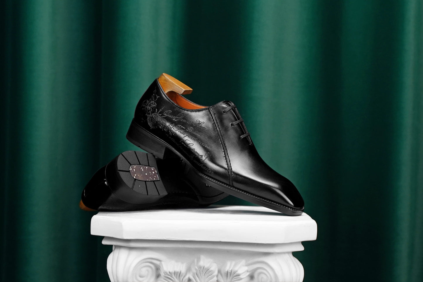 A pair of polished black leather dress shoes displayed on a white decorative pedestal against a dark green draped curtain background.