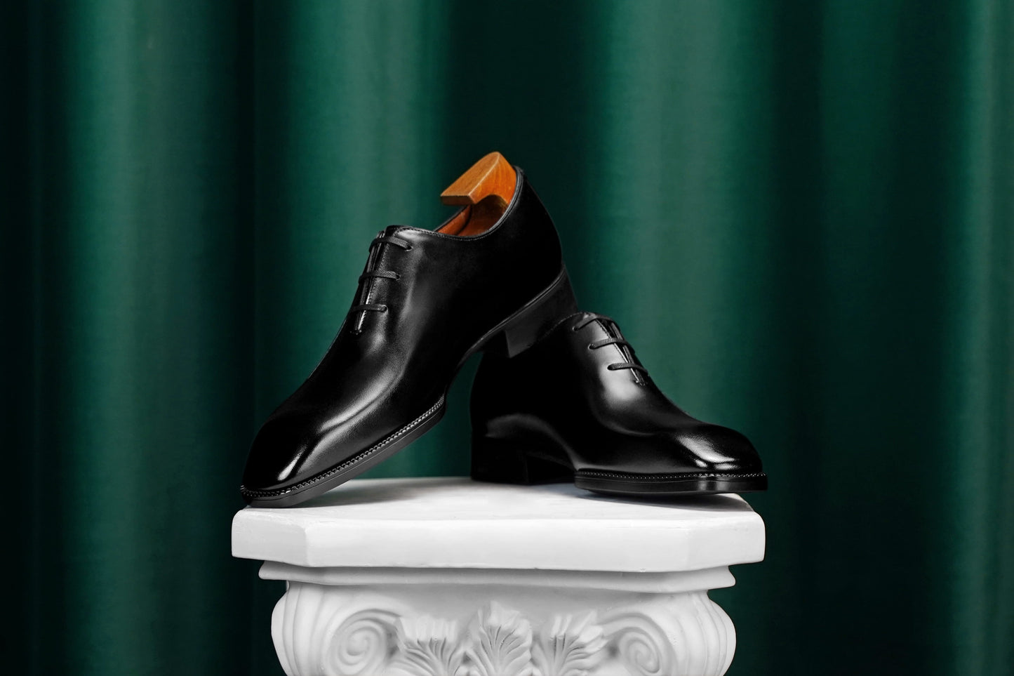 A pair of polished black leather oxford dress shoes displayed on a white decorative pedestal against a dark green draped curtain background.