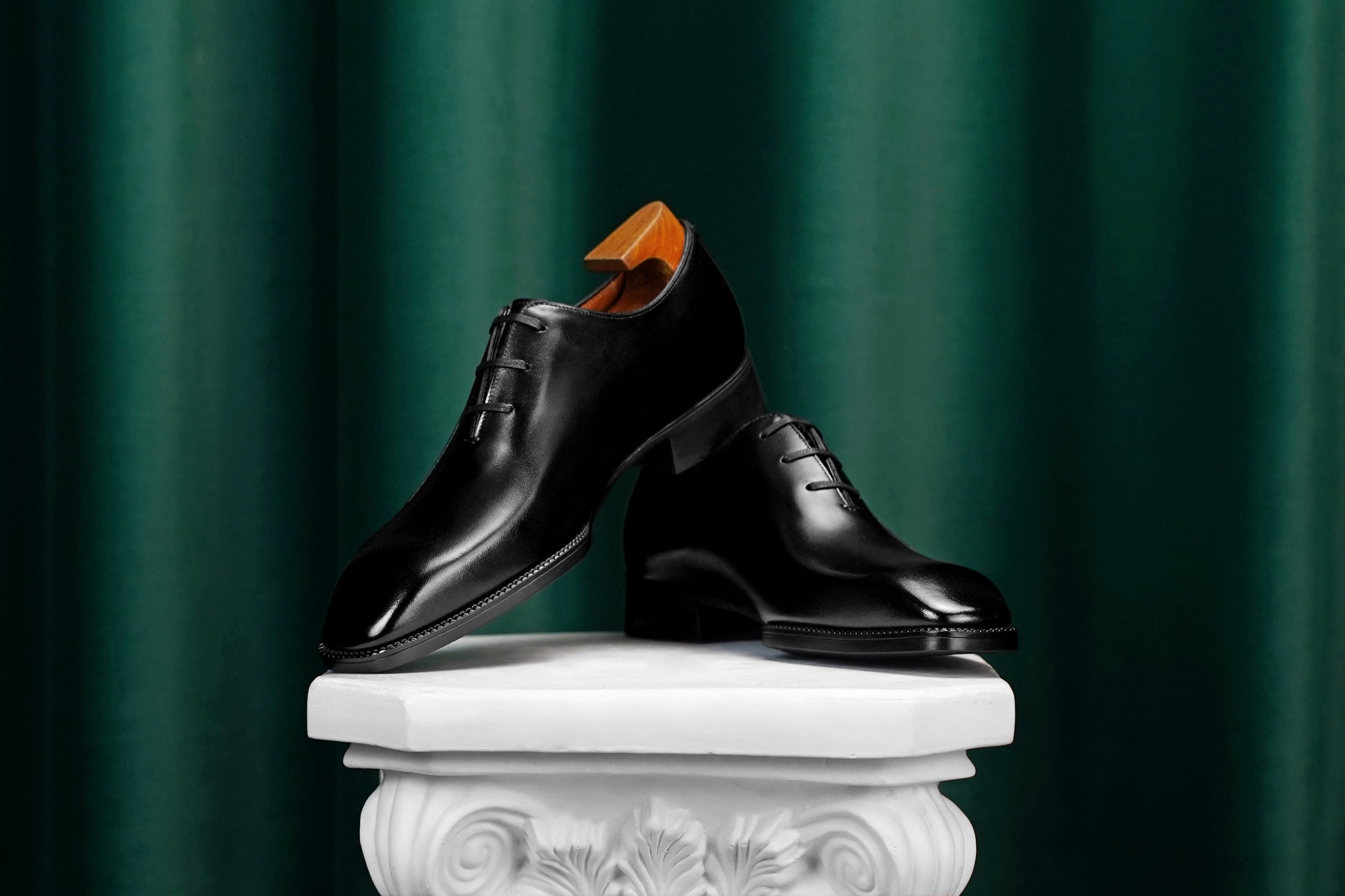 A pair of polished black leather oxford dress shoes displayed on a white decorative pedestal against a dark green draped curtain background.
