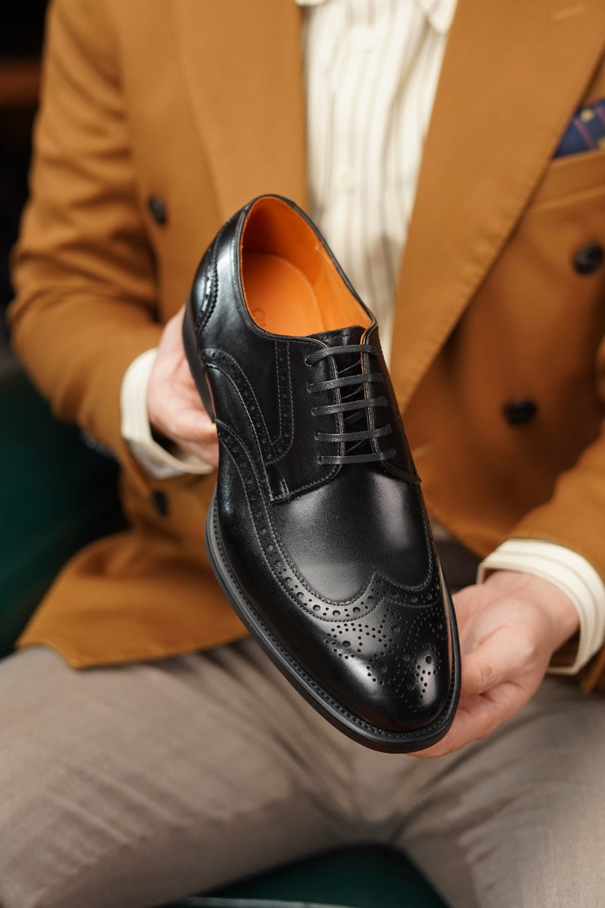 A man in a brown blazer and beige trousers holds up a black wingtip brogue oxford shoe with a contrasting orange interior.
