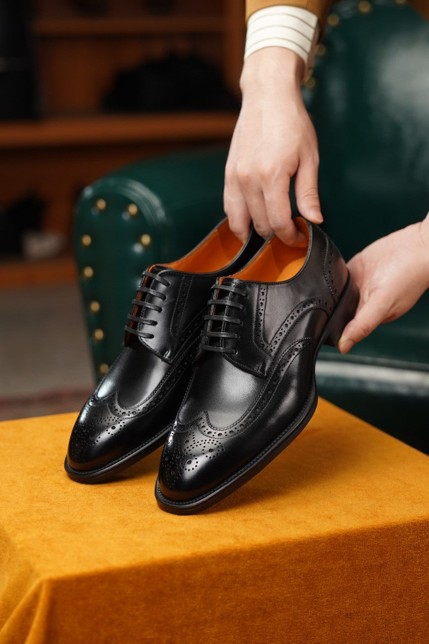 A pair of black leather wingtip oxford shoes with brogue detailing, held by a person's hands over an orange fabric-covered surface in a warm indoor setting.