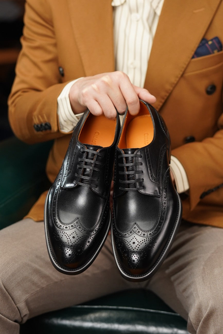 A person in a tan blazer and beige trousers holds a pair of black brogue oxford shoes with orange lining against a dark green leather seat.
