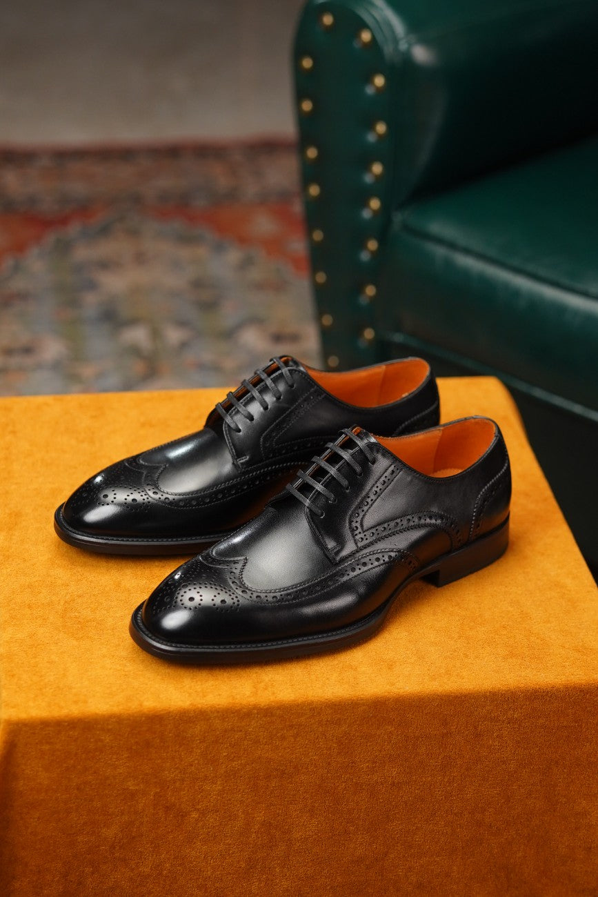 A pair of black leather wingtip oxford shoes with brogue detailing, displayed on a textured orange block with a green leather armchair in the background.
