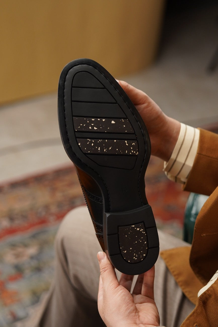 A close-up of a person holding the outsole of a brown leather shoe to show the tread pattern and rubber heel piece. The person is wearing a brown jacket and beige trousers, with a patterned rug and wooden wall in the background.
