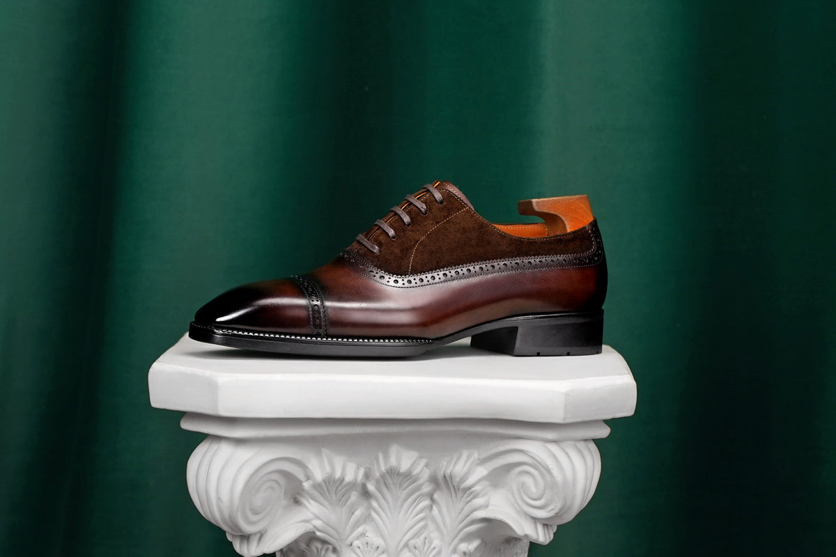 A single men's brogue oxford shoe in dark brown and cognac brown leather, displayed on a white decorative pedestal against a dark green draped background.
