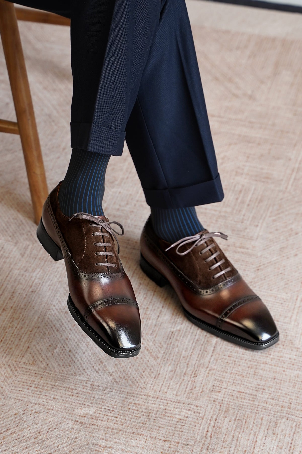 A close-up of a person's legs wearing dark navy trousers, dark blue striped socks, and brown leather cap-toe brogue oxford shoes with dark soles, standing on a textured beige carpet.
