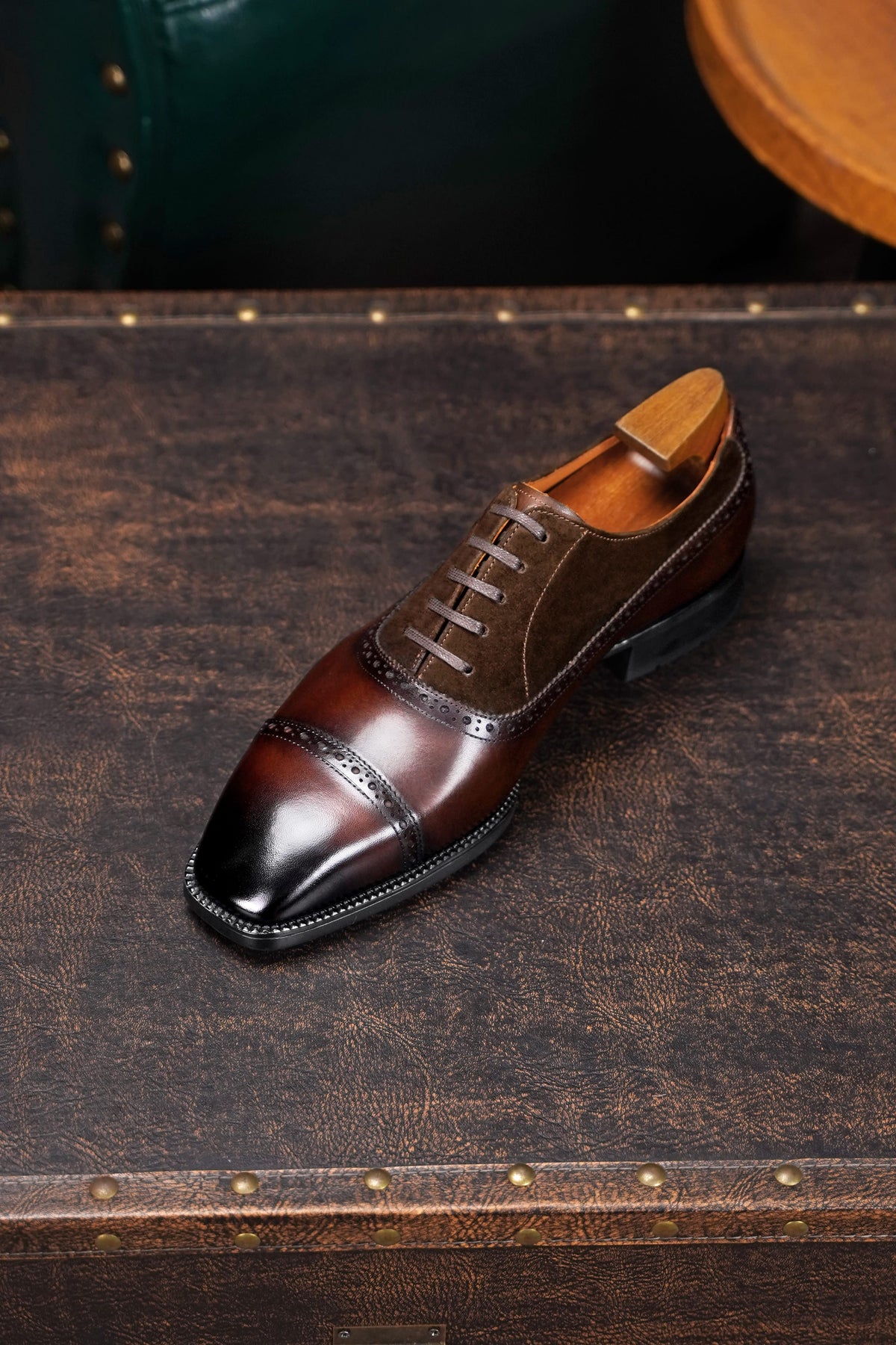 A dark brown brogue oxford shoe with a glossy cap toe and black sole, displayed on a vintage brown leather suitcase.