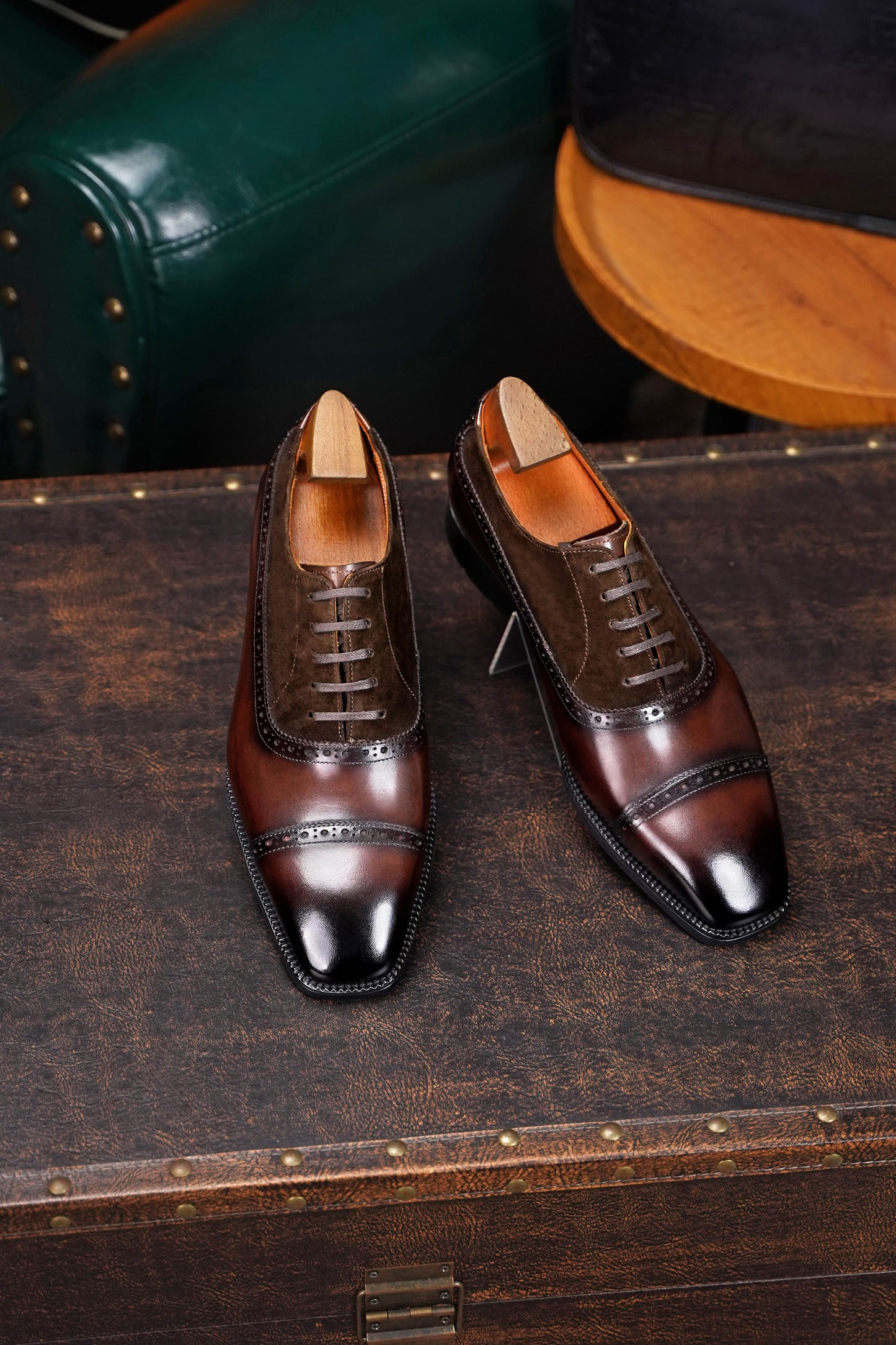 Pair of brown leather dress shoes on a wooden surface with a vintage suitcase in the background.