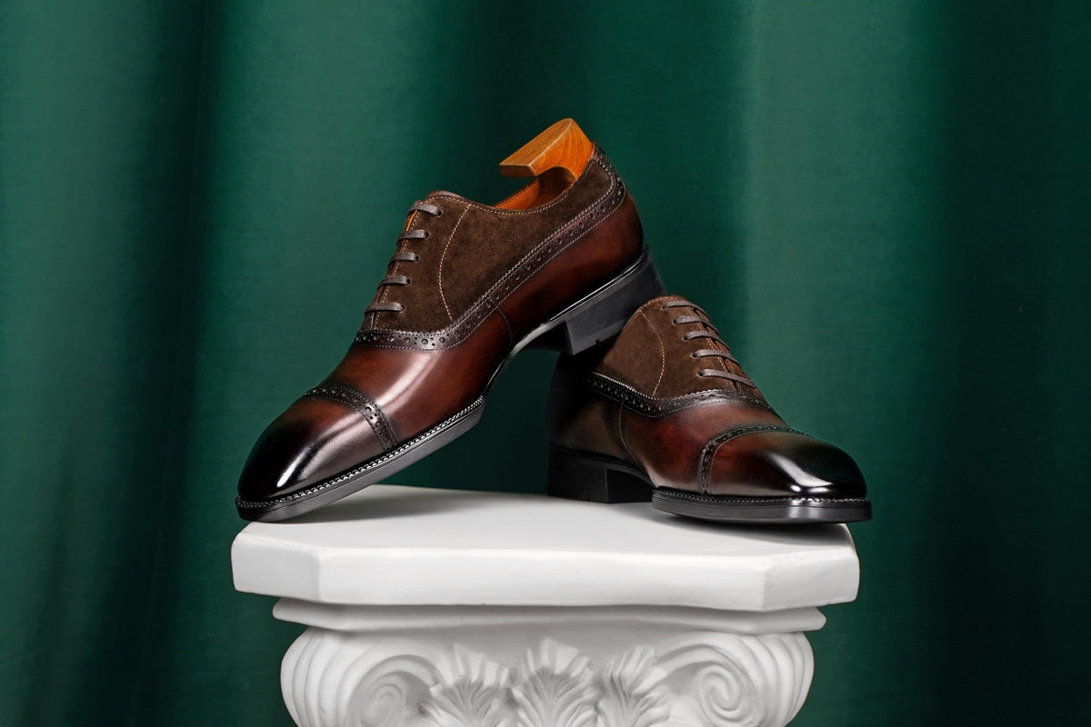 A pair of dark brown leather cap-toe oxford shoes with a mix of glossy patent leather and suede panels, displayed on a white decorative pedestal against a dark green draped background.