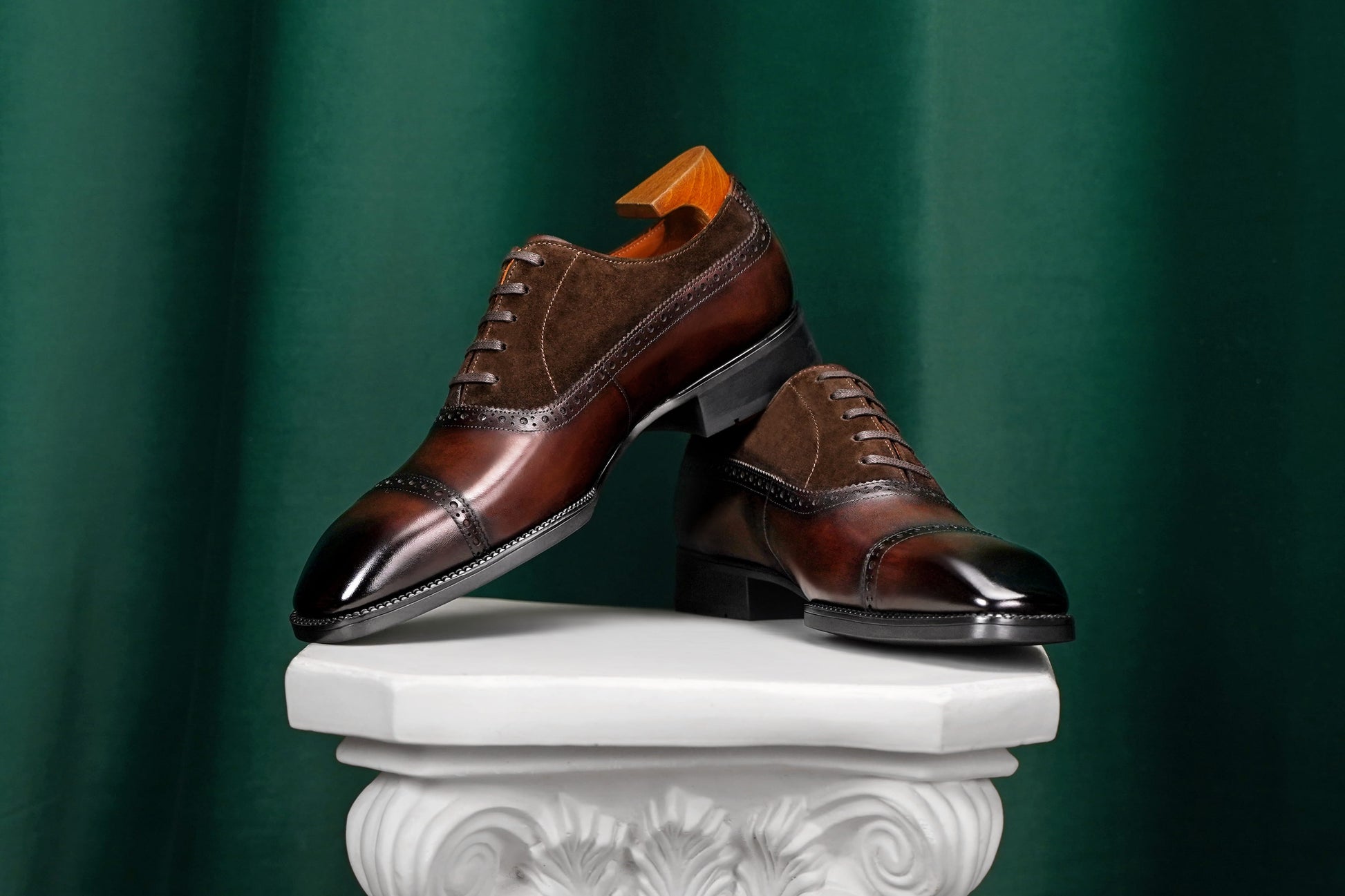 A pair of dark brown leather cap-toe oxford shoes with a mix of glossy patent leather and suede panels, displayed on a white decorative pedestal against a dark green draped background.