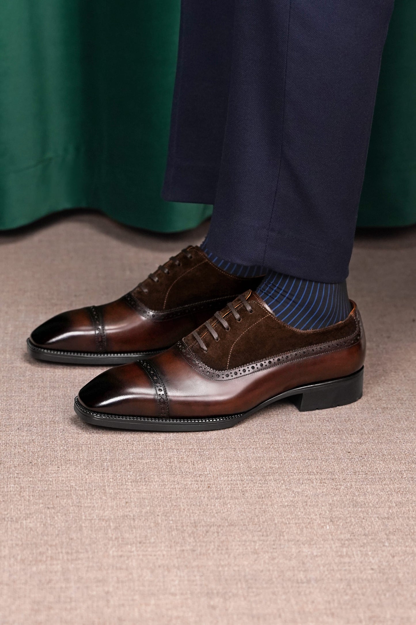 A pair of men's brown leather cap-toe oxford shoes with brogue detailing, styled with navy trousers and blue striped socks on a textured beige carpet.