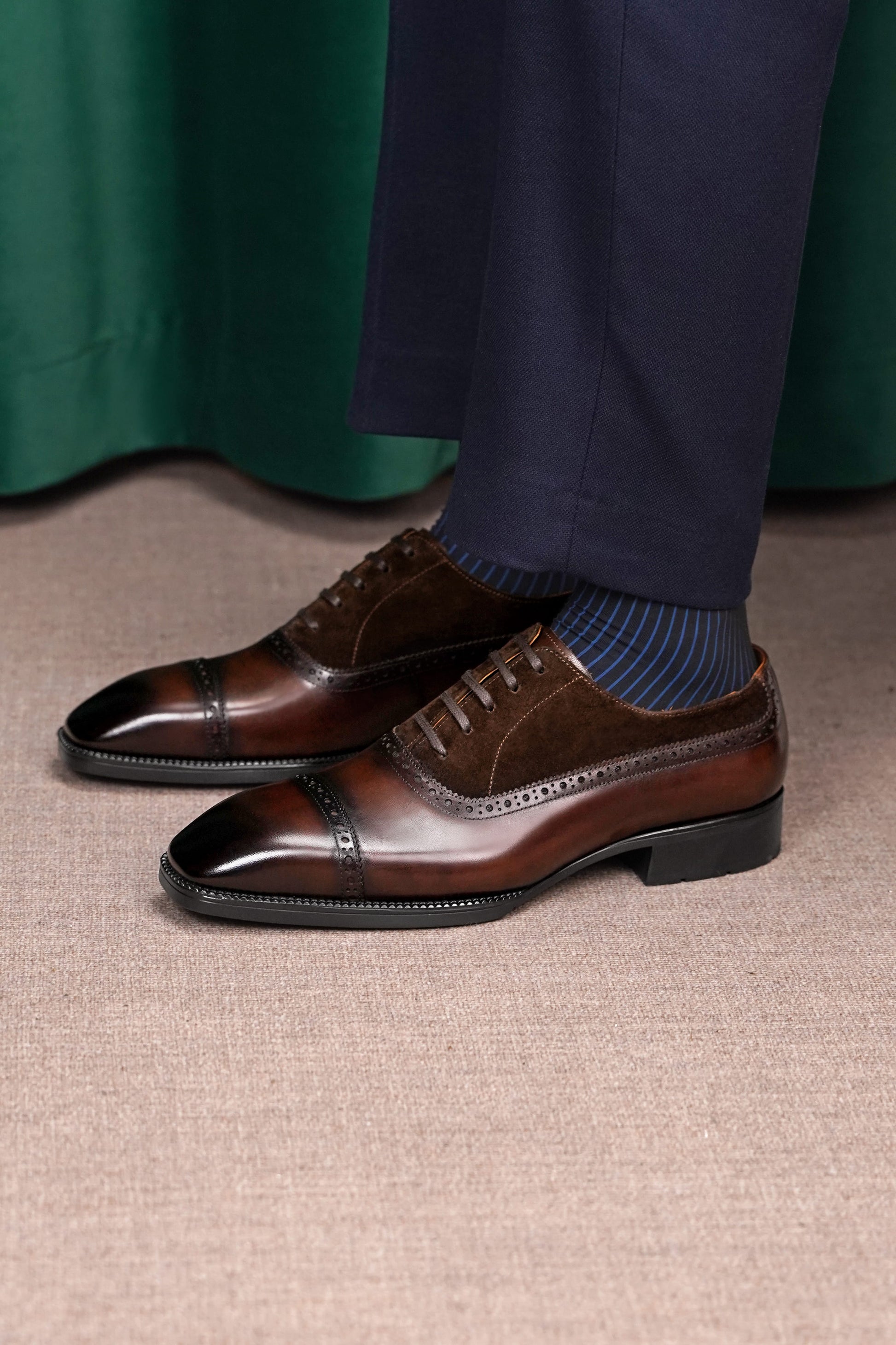A pair of men's brown leather cap-toe oxford shoes with brogue detailing, styled with navy trousers and blue striped socks on a textured beige carpet.
