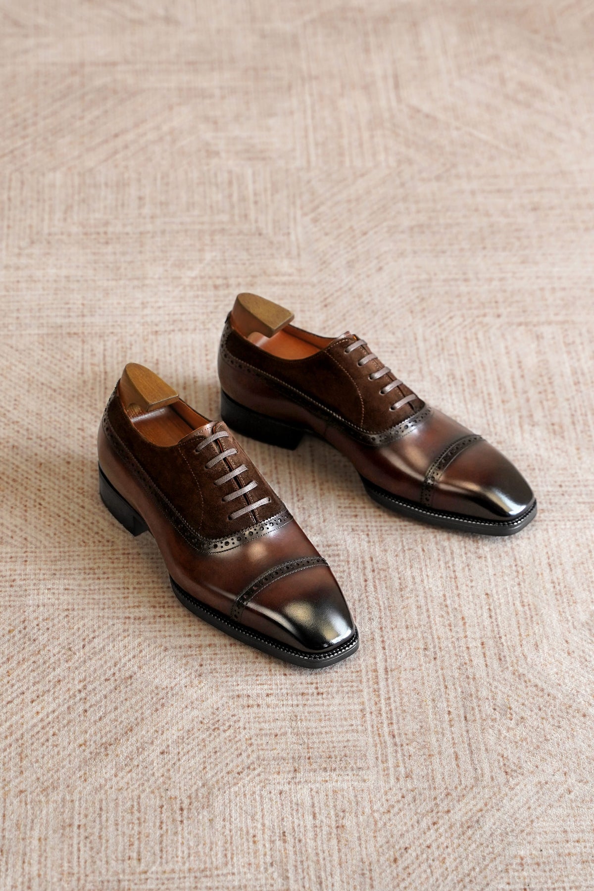 A pair of dark brown leather cap-toe oxford shoes with brogue detailing, displayed on a textured beige woven rug.