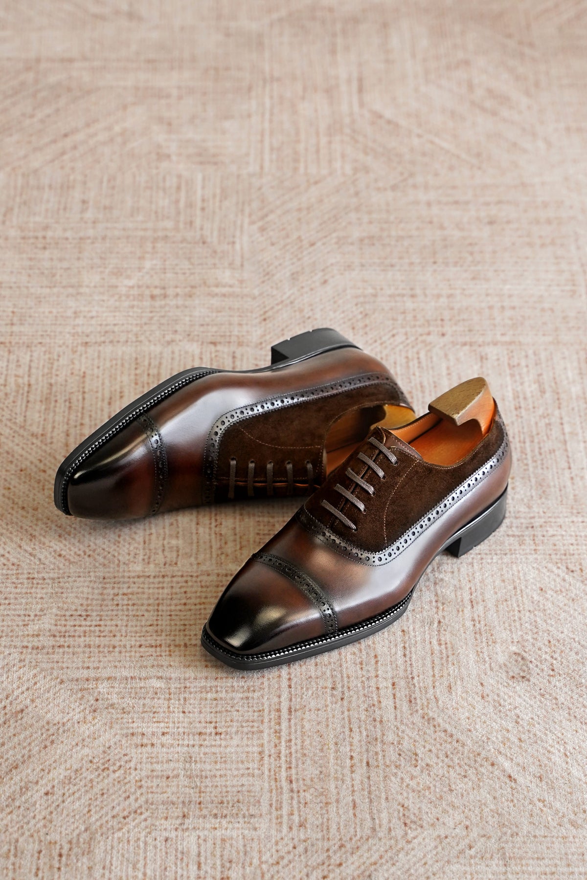 A pair of dark brown brogue oxford shoes with a mix of patent leather and suede panels, displayed on a textured beige woven rug.