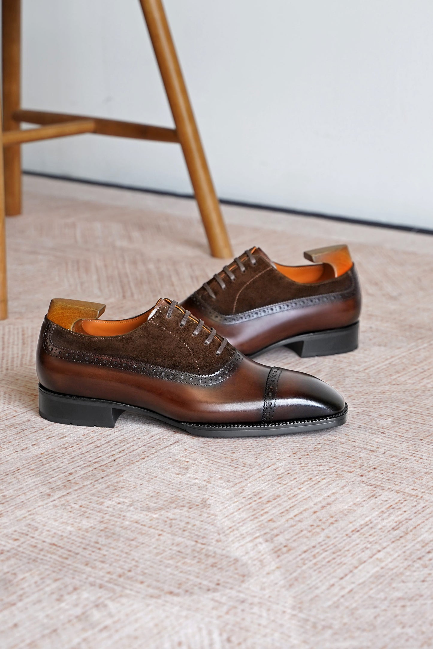 A pair of men's brown leather oxford shoes with a cap toe and brogue detailing, displayed on a textured beige carpet with a wooden chair leg visible in the background.