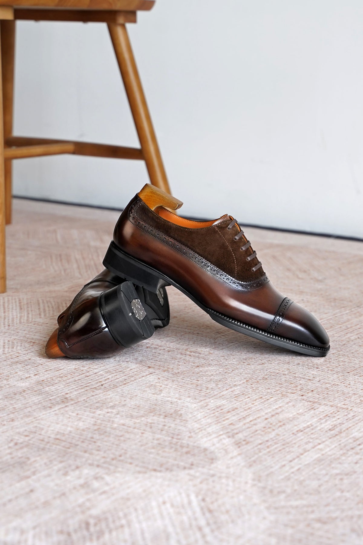 A pair of men's brown leather wingtip oxford shoes with brogue detailing, displayed on a textured beige rug with a wooden stool leg visible in the background.