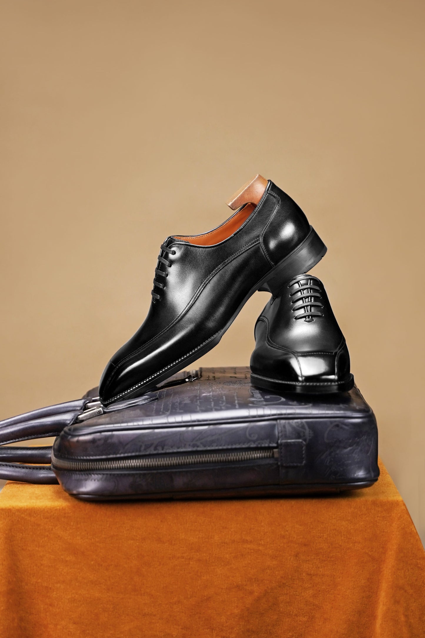 A pair of black leather dress shoes displayed on top of a black leather briefcase, set against a warm beige background with an orange fabric-covered pedestal.