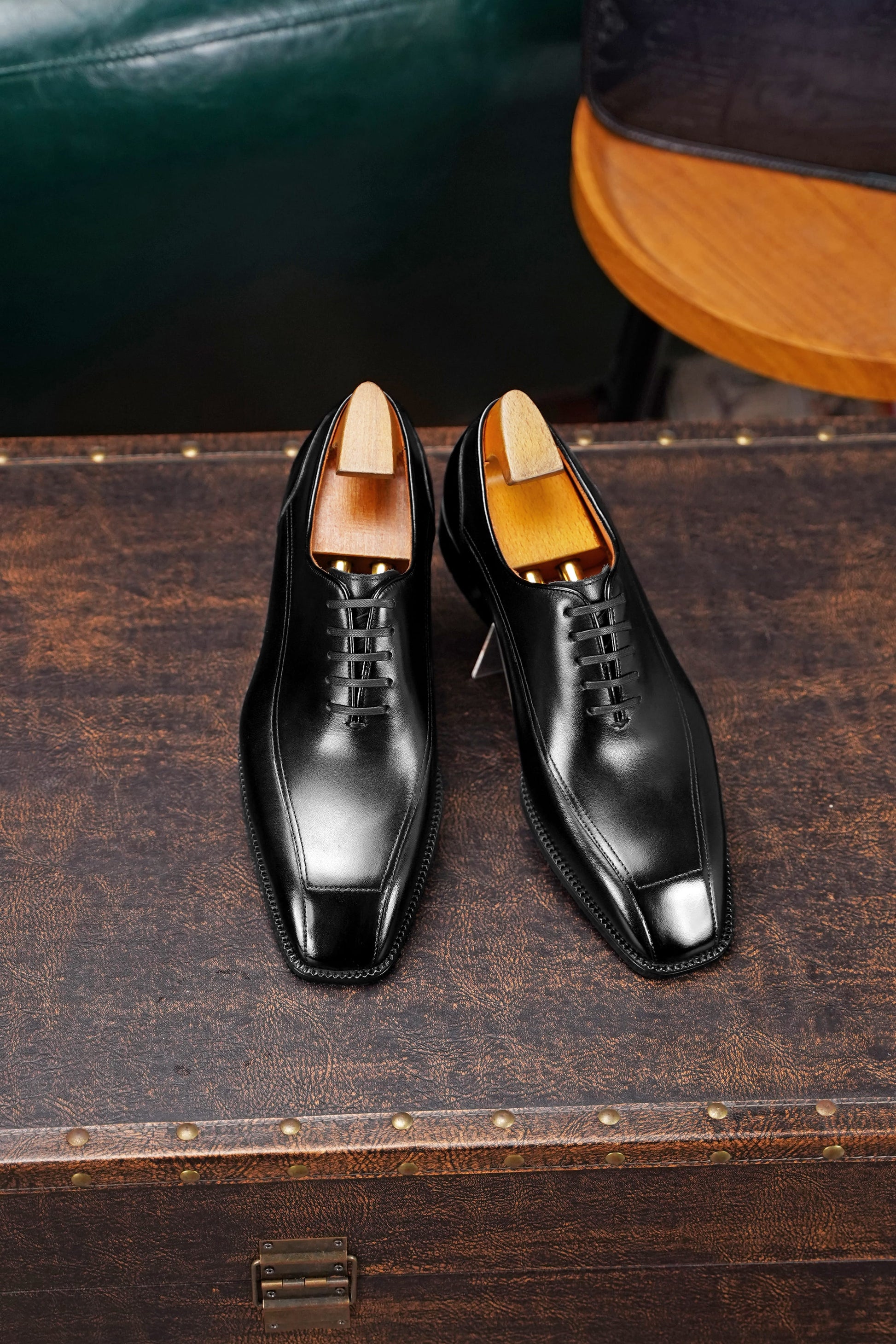 A pair of black leather oxford dress shoes with wooden shoe trees, displayed on a dark brown wooden trunk.