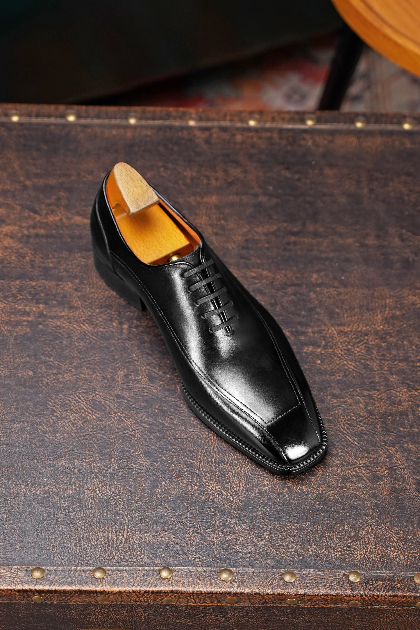 A single black leather dress shoe with a square toe and lace-up front, displayed on a dark brown textured leather surface with a wooden shoe tree visible inside.