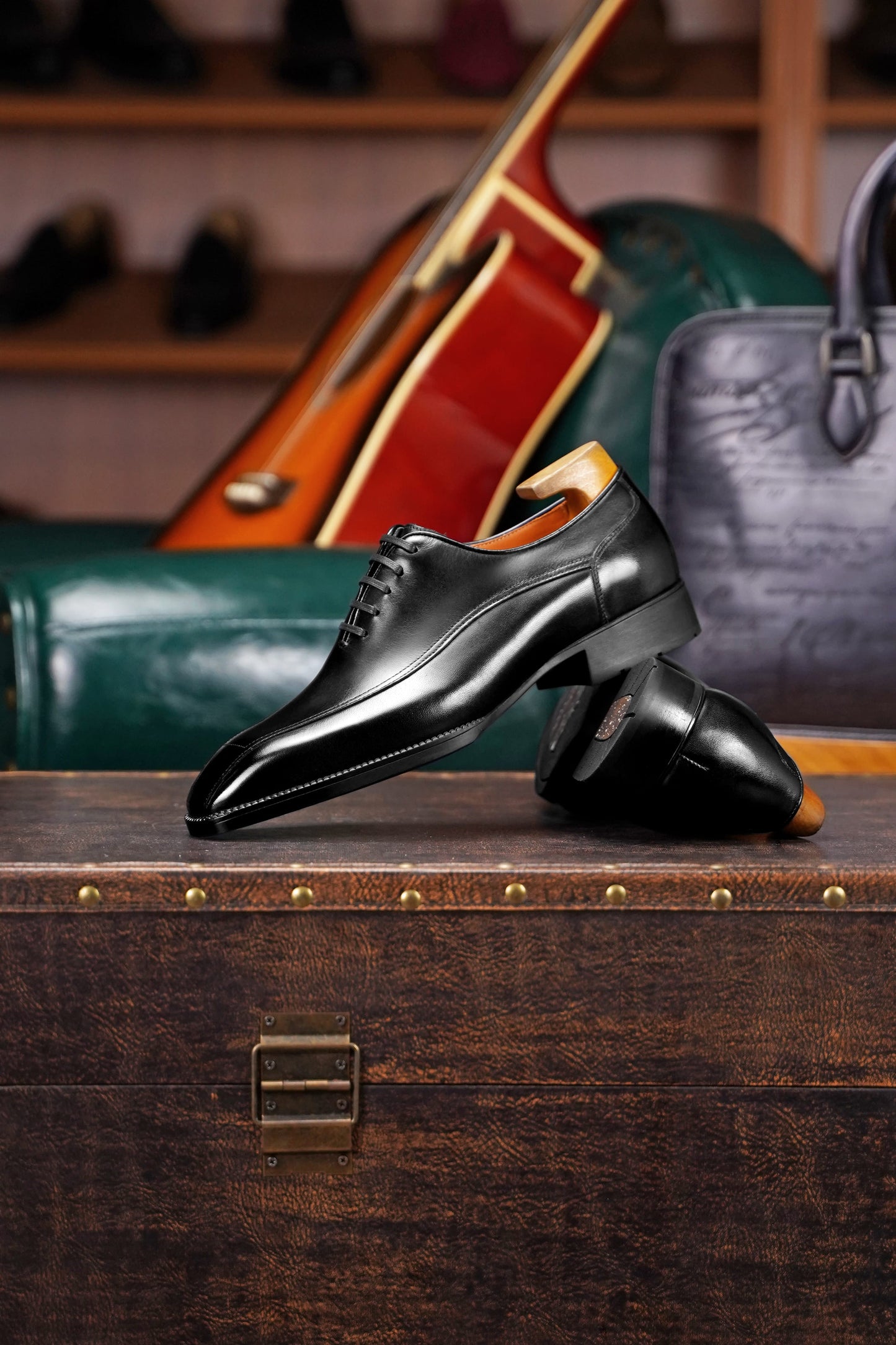 A pair of polished black leather dress shoes with a low block heel and lace-up front, displayed on a rustic wooden trunk with a cello and leather handbag in the background.
