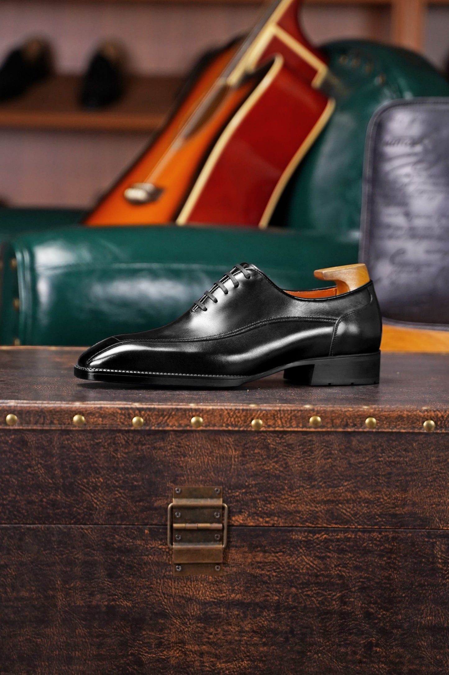 A single black leather oxford dress shoe with a wingtip toe and lace-up front, displayed on a vintage wooden trunk in a styled interior setting with a green leather chair and guitar in the background.
