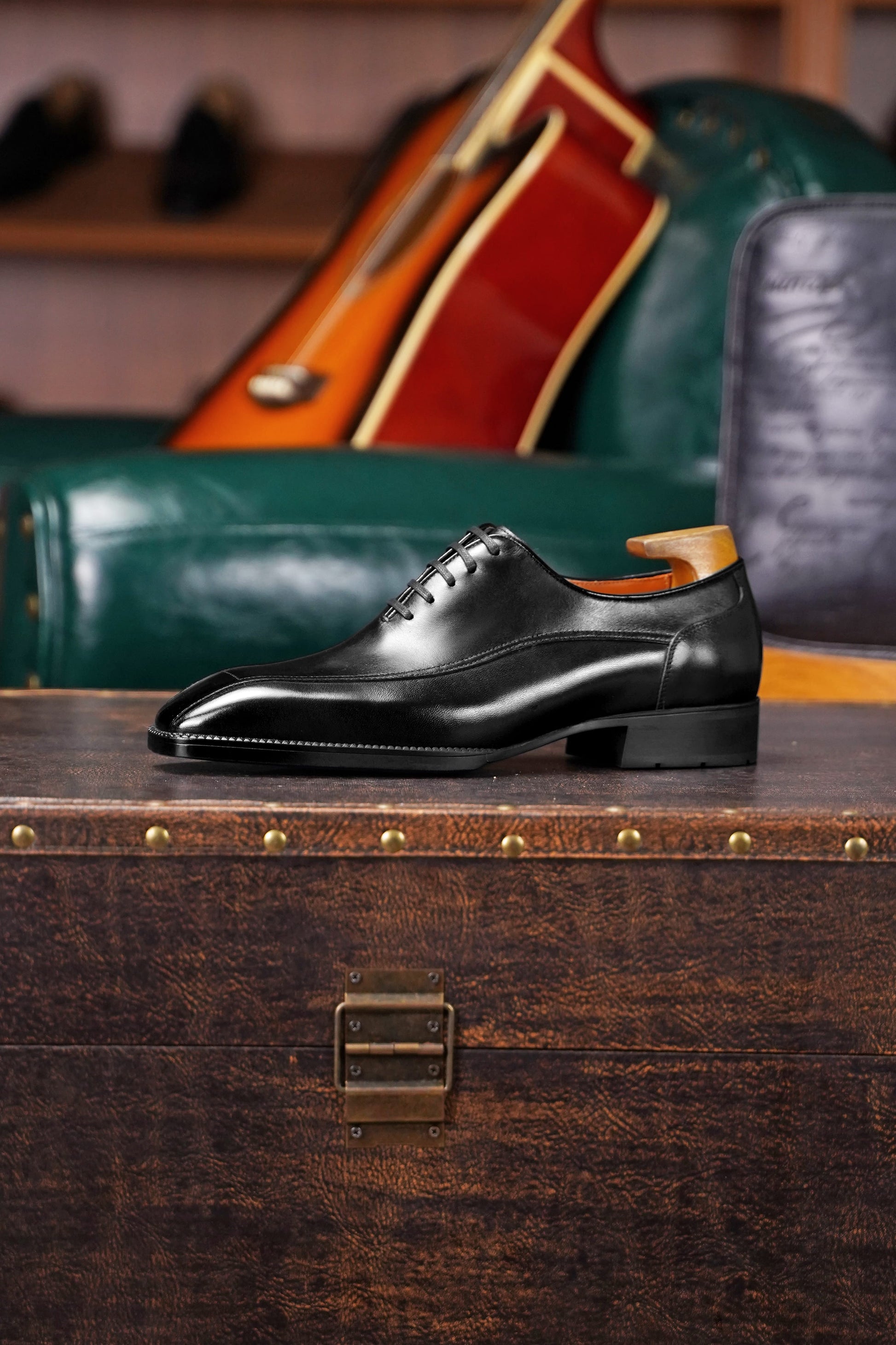 A single black leather oxford dress shoe with a wingtip toe and lace-up front, displayed on a vintage wooden trunk in a styled interior setting with a green leather chair and guitar in the background.