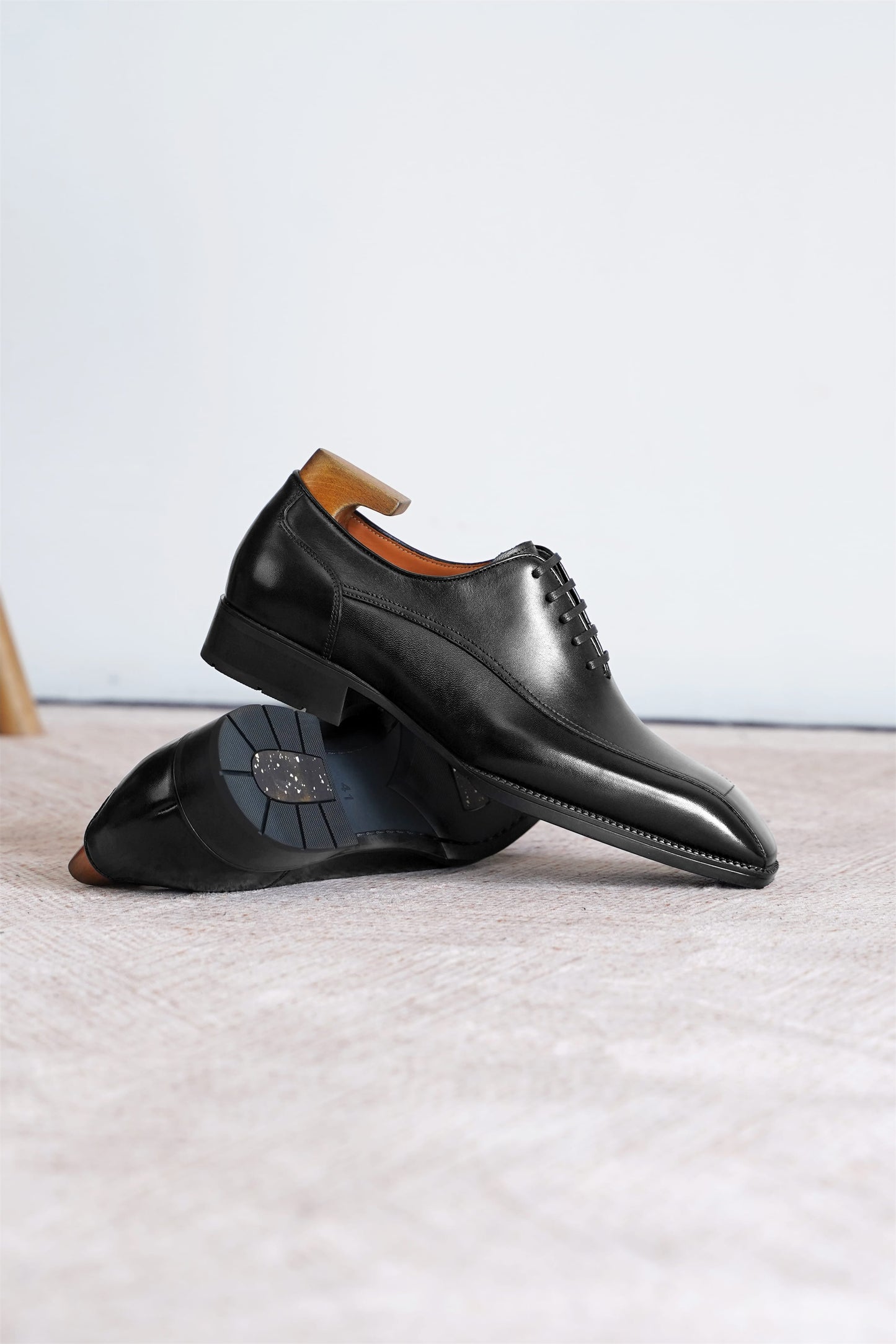 A pair of black leather dress shoes with a pointed toe and lace-up closure, displayed on a light beige textured surface against a white background.