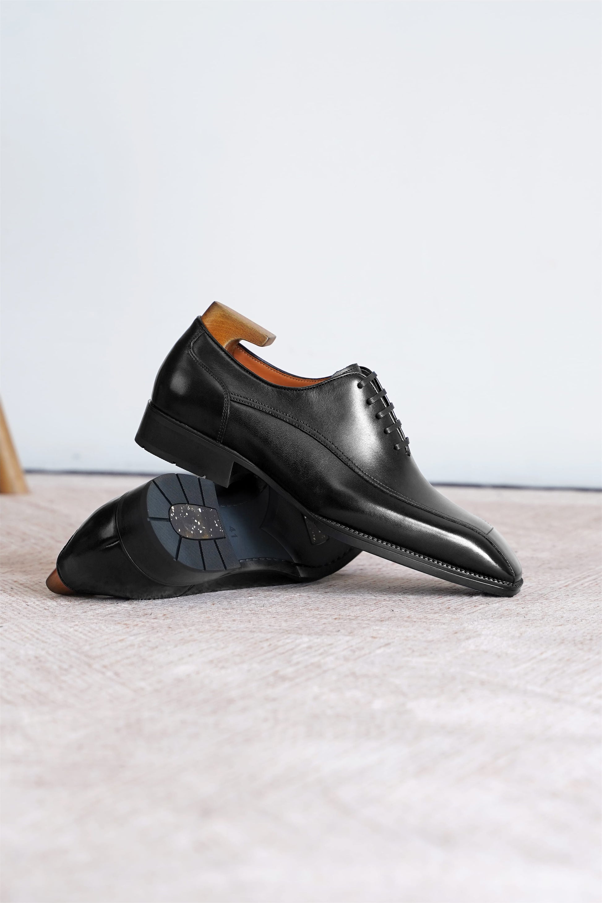 A pair of black leather dress shoes with a pointed toe and lace-up closure, displayed on a light beige textured surface against a white background.