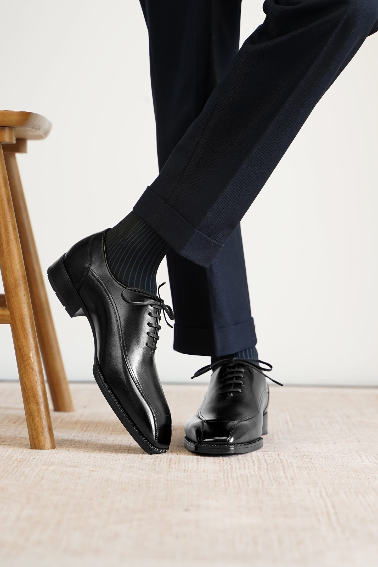 A close-up of a person's legs wearing black leather oxford dress shoes, dark ribbed socks, and navy trousers, standing on a light beige textured floor next to a wooden stool.