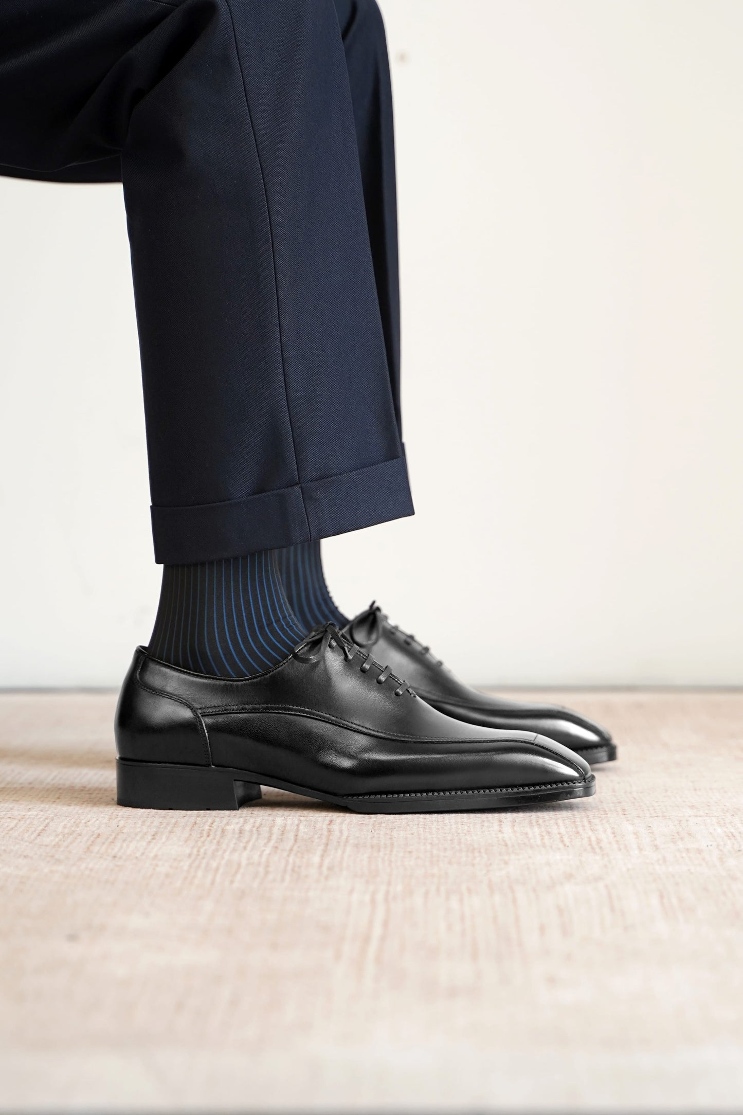 A close-up of a person's legs wearing dark navy trousers, blue and black striped socks, and black leather oxford dress shoes on a light beige textured floor against a neutral off-white background.