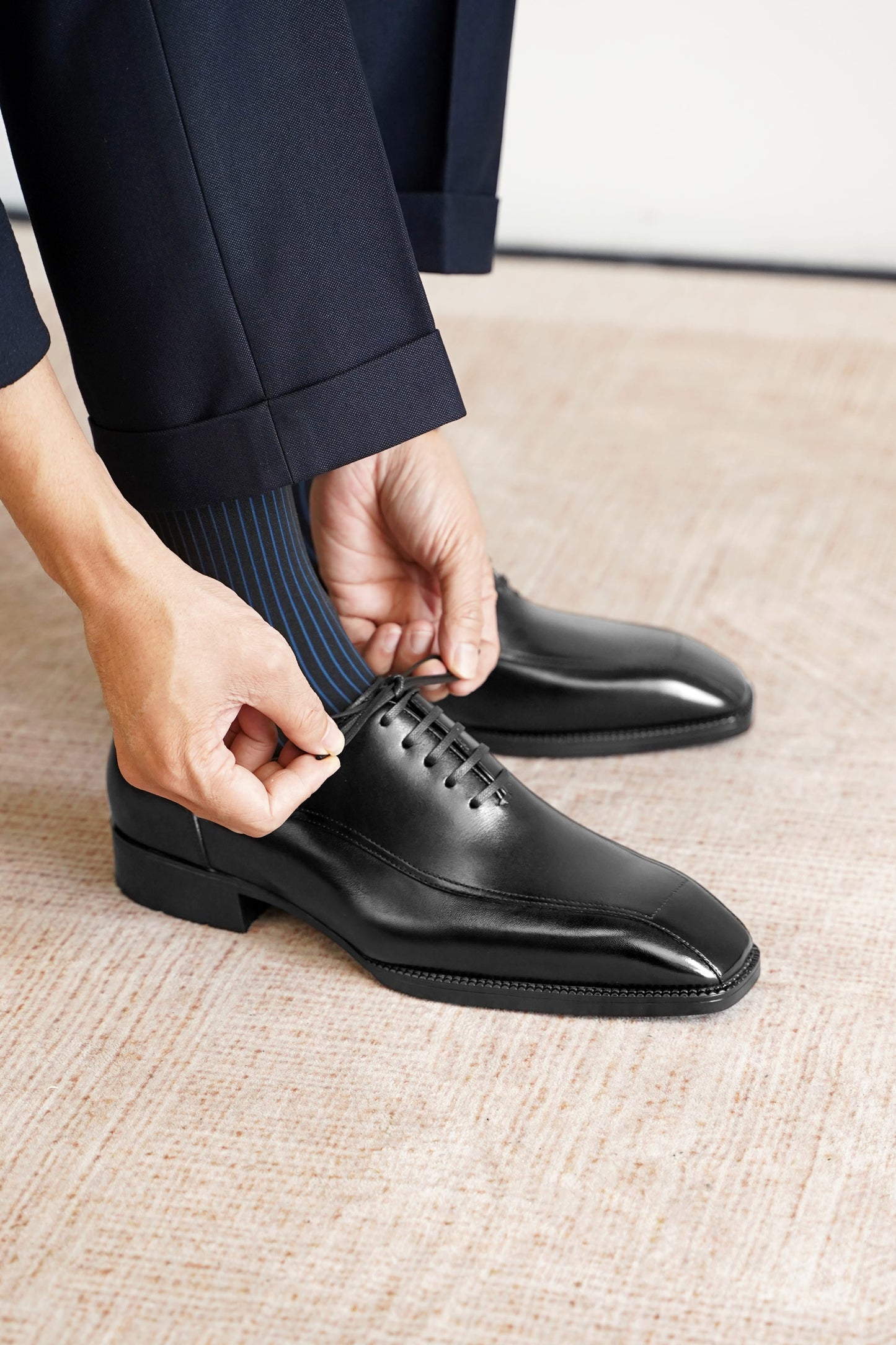 A close-up of a person's hands adjusting a dark navy dress sock on their feet, which are wearing polished black oxford shoes. The person is also wearing dark navy trousers.