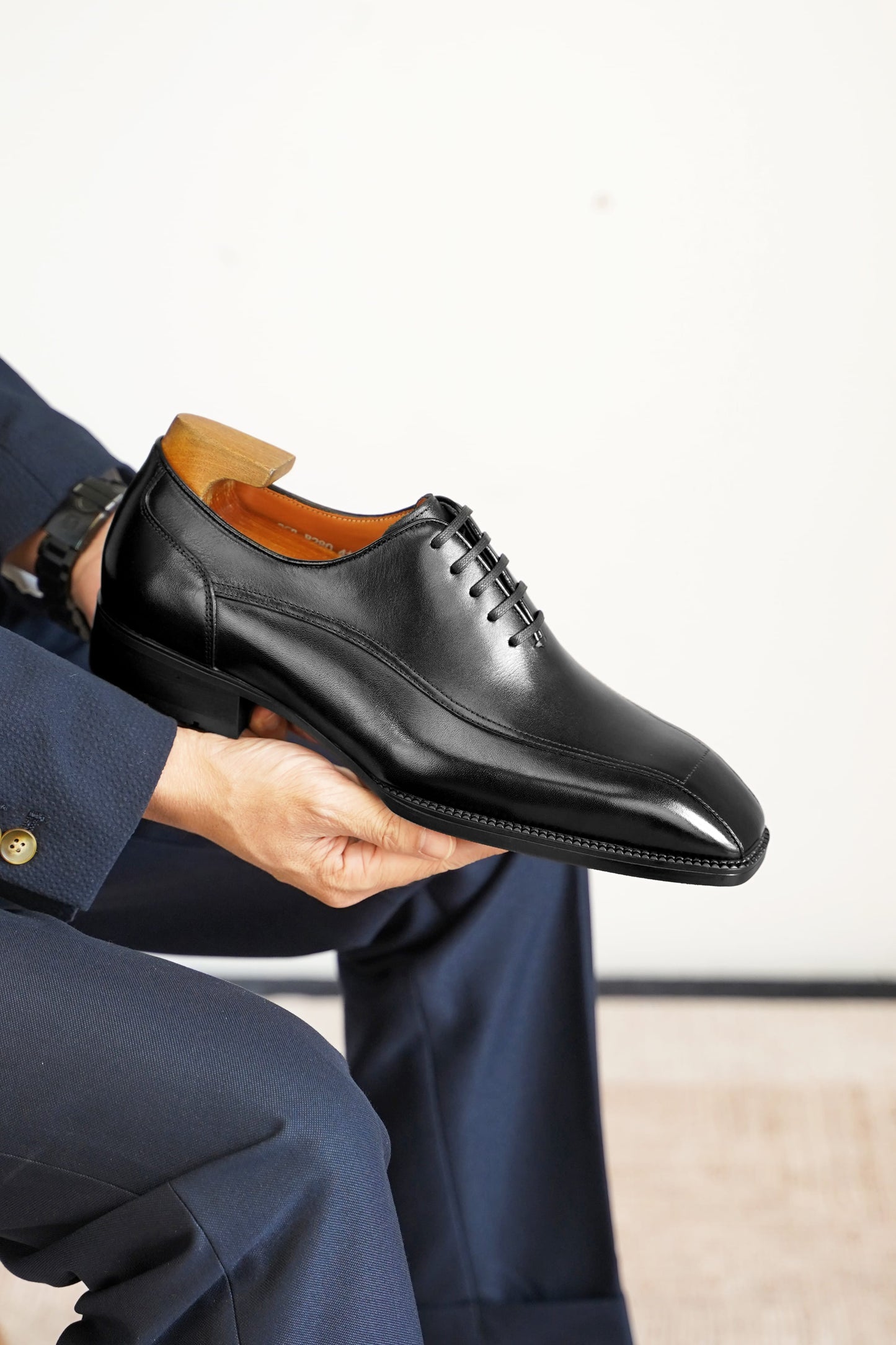 A man in a navy blue suit holds up a black leather oxford dress shoe with a brown wooden shoe tree insert. The shoe is shown from a side angle, highlighting its smooth leather finish, black laces, and the warm brown interior lining.
