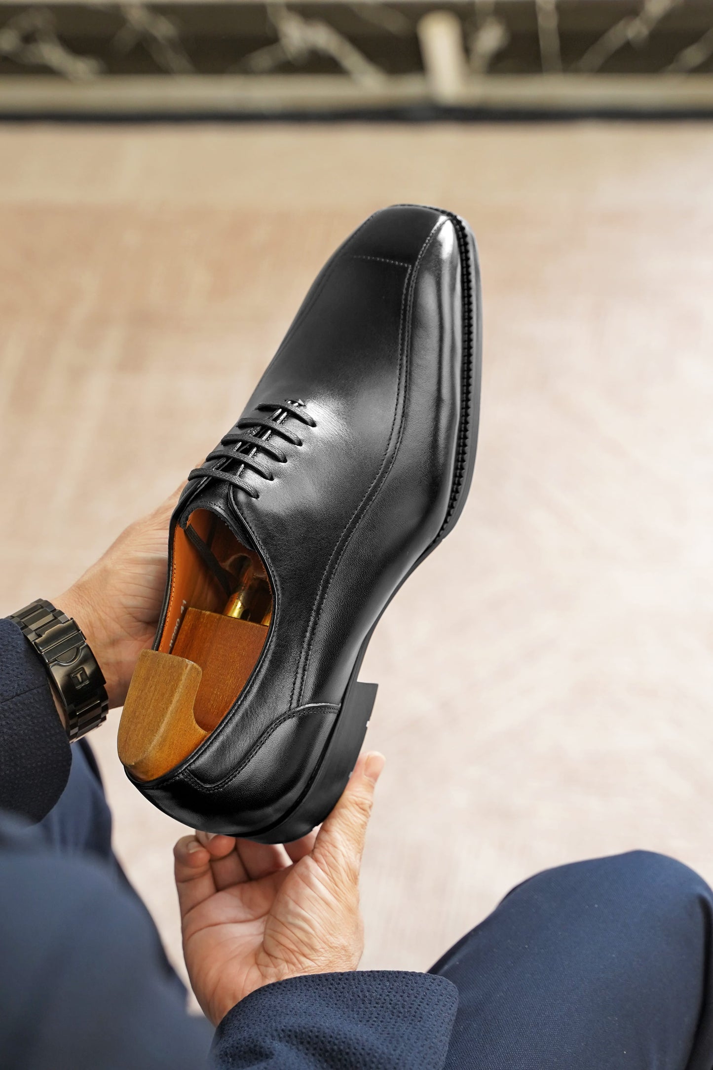 A black leather dress shoe with a wooden shoe tree insert, held by hands wearing a navy blazer and a black watch, against a blurred beige background.