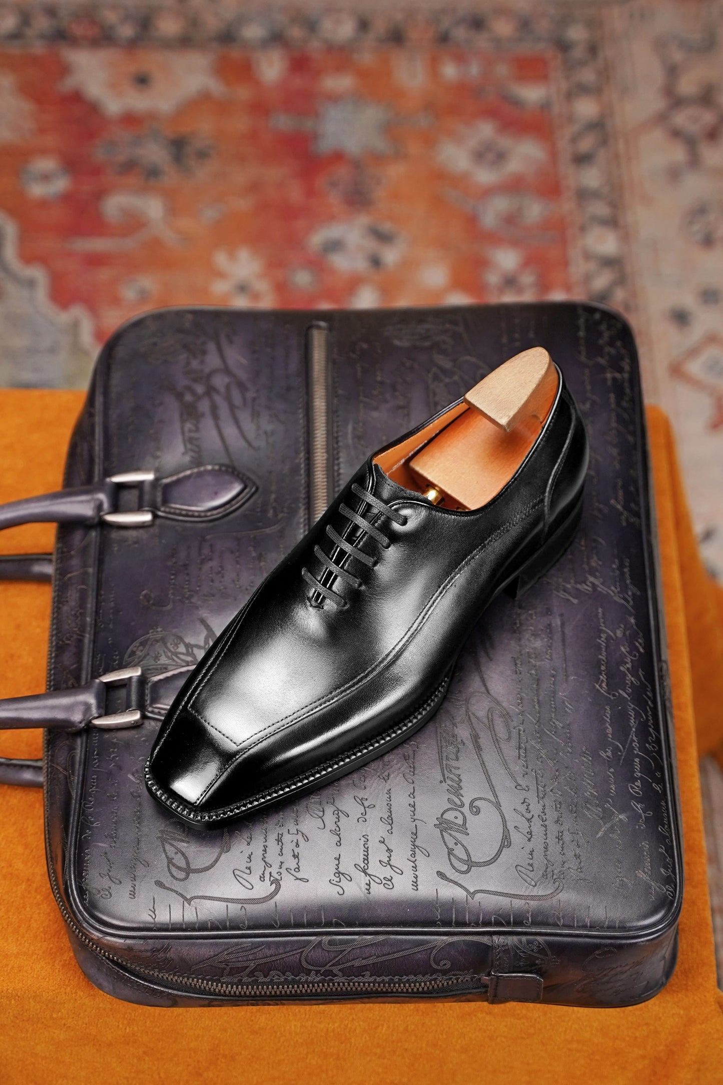 A black leather dress shoe with a square toe and lace-up front, displayed on top of a black embossed leather briefcase or bag, set against a patterned rug and orange fabric surface.