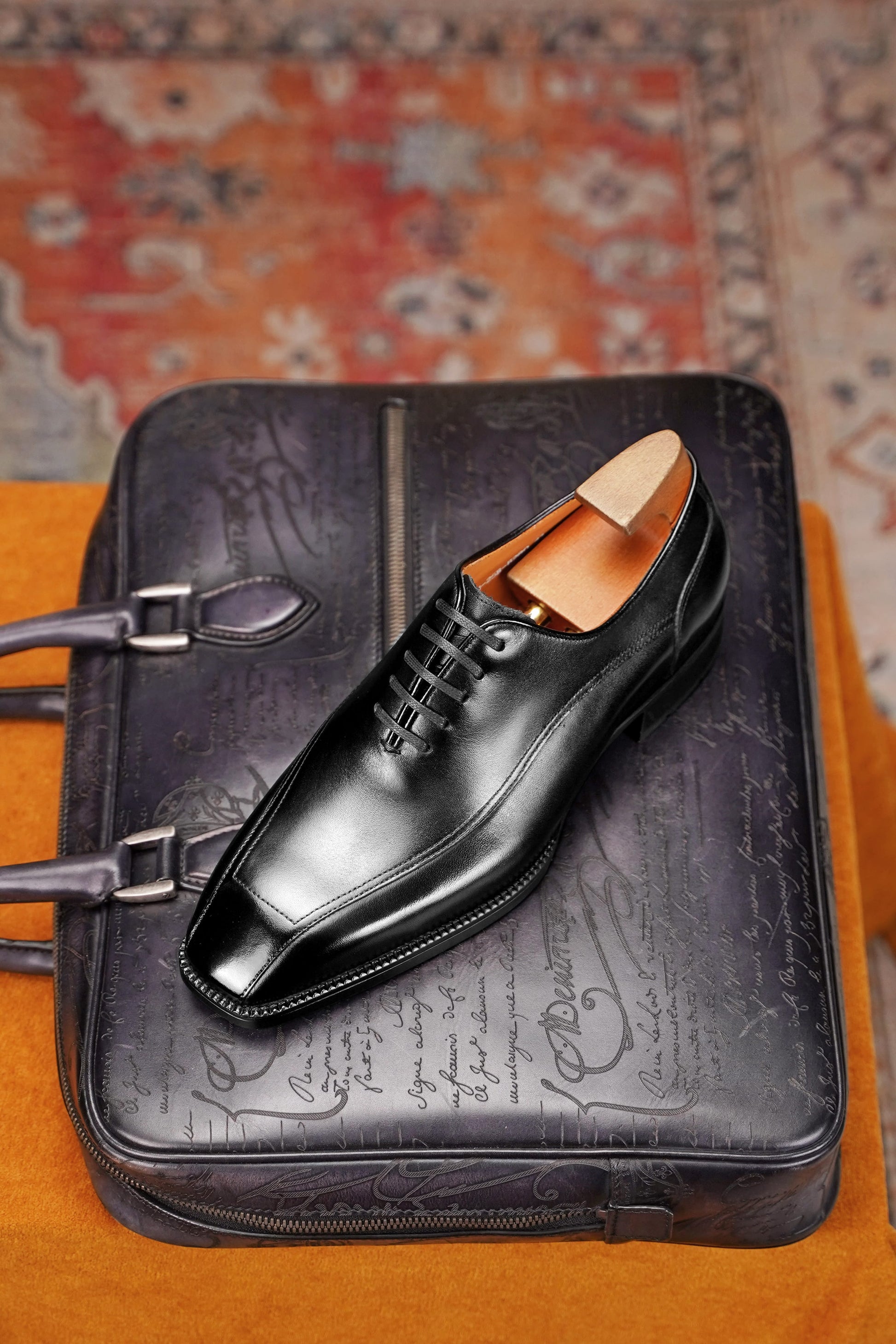A black leather dress shoe with a square toe and lace-up front, displayed on top of a black embossed leather briefcase or bag, set against a patterned rug and orange fabric surface.
