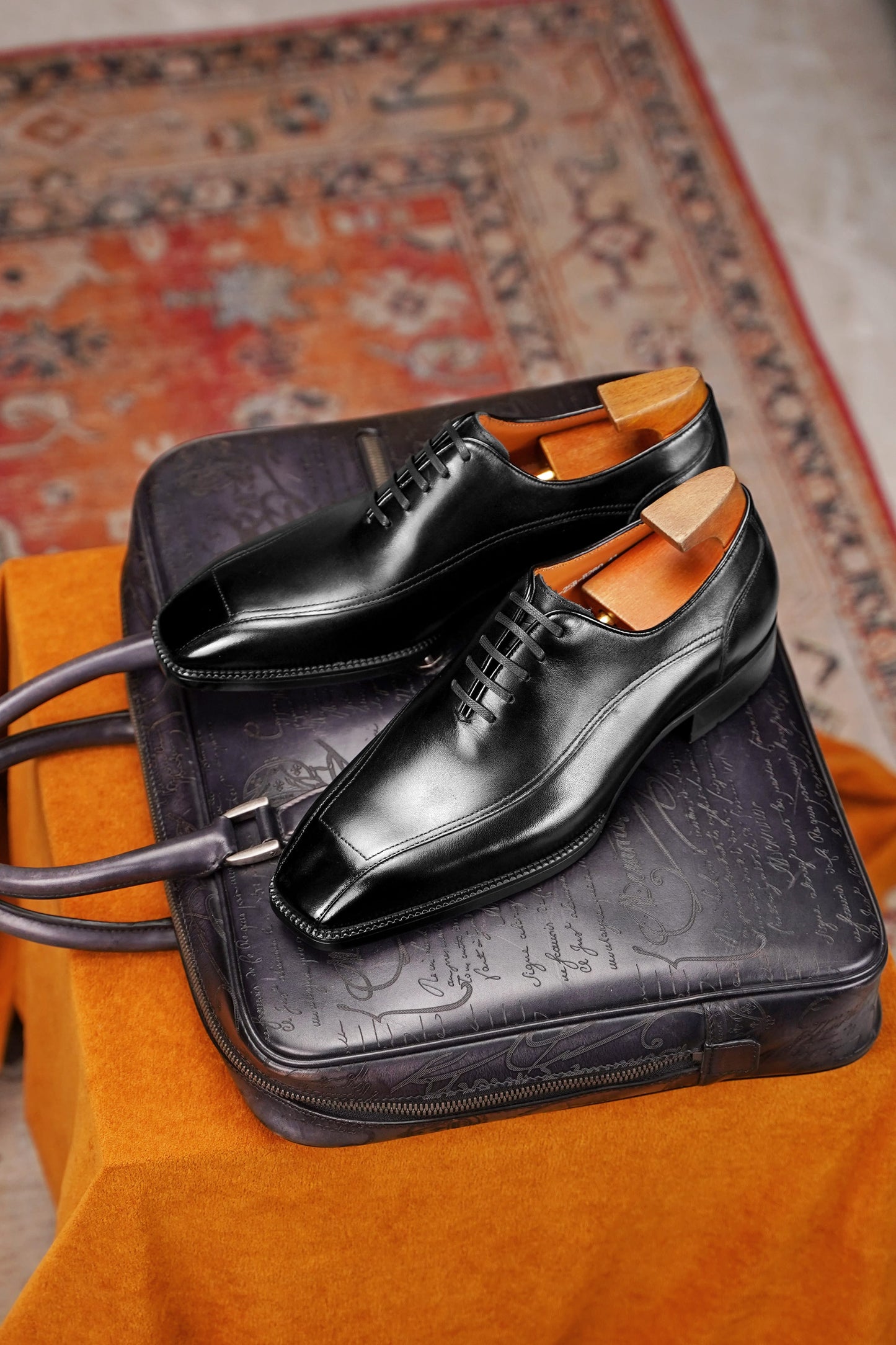 A pair of black leather oxford dress shoes with light wood shoe trees inside, displayed on top of a dark embossed leather briefcase or suitcase, set against an orange fabric surface with a patterned rug in the background.