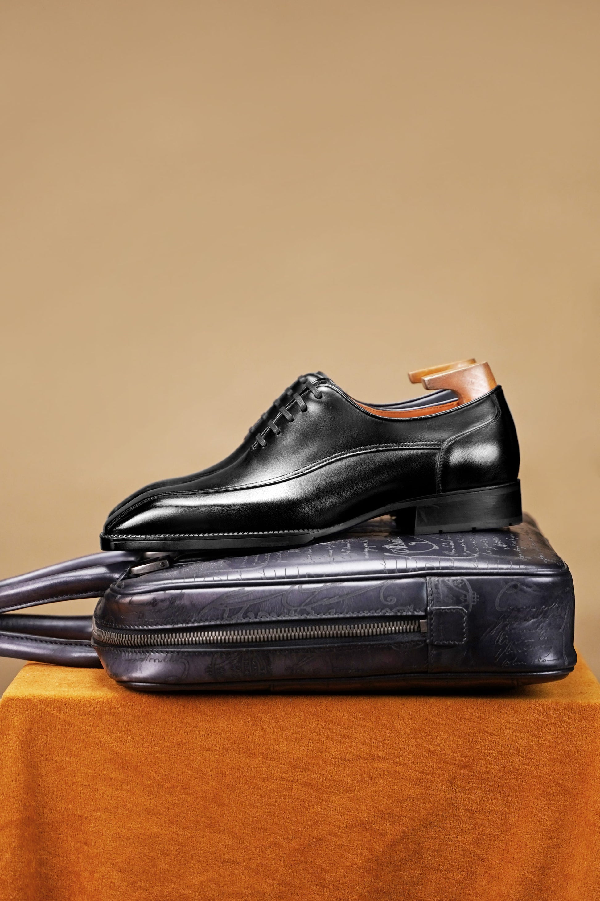 A black leather oxford dress shoe with a brown wooden shoe tree inserted, displayed on top of a dark textured leather zippered case against a warm beige background.