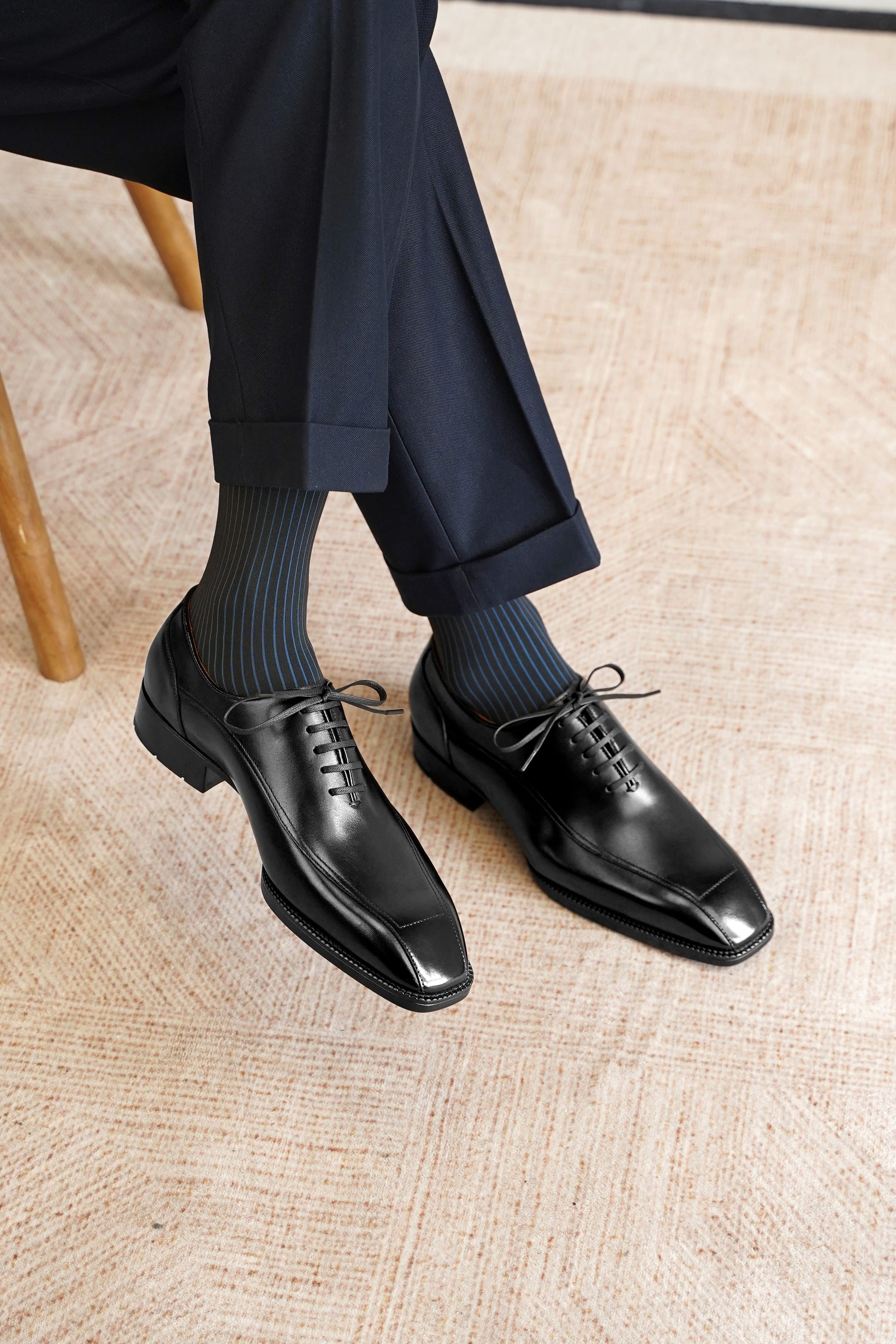 A person seated on a wooden chair wearing black leather oxford dress shoes with dark ribbed socks and navy trousers, photographed on a light beige woven rug.