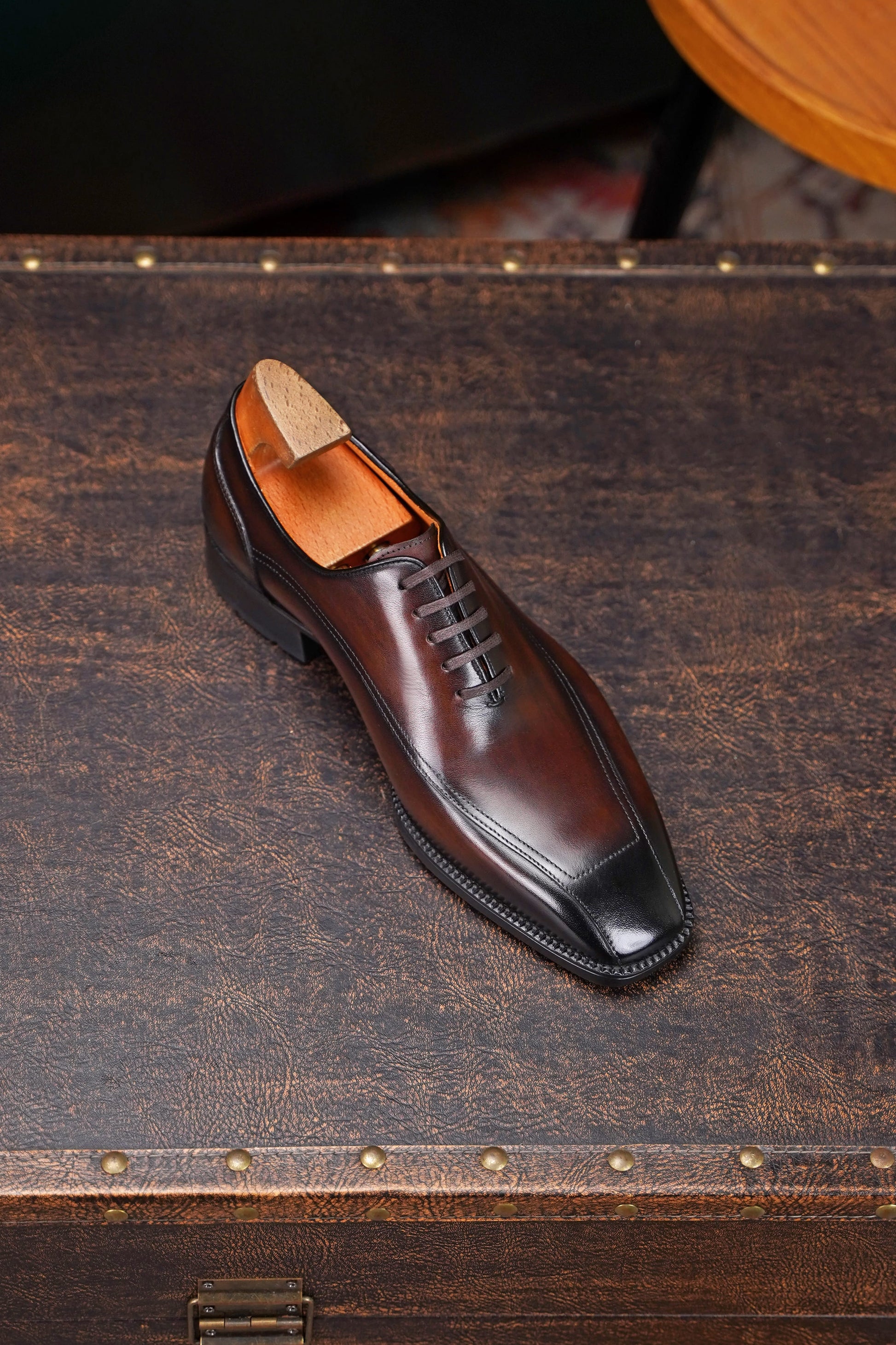 A dark brown leather dress shoe with a black cap toe and laces, shown with a wooden shoe tree insert, resting on a rustic dark brown leather suitcase.