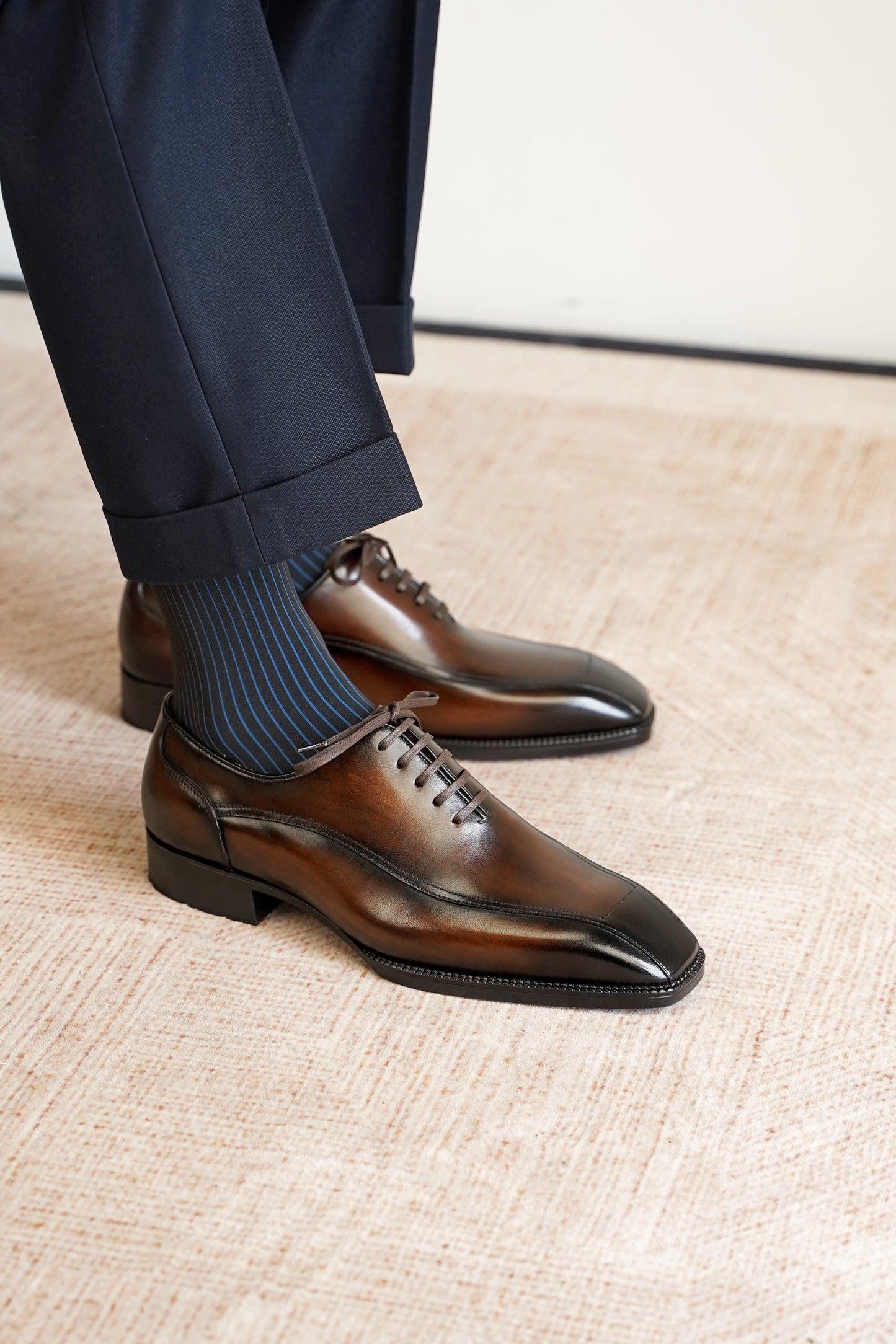 A close-up of a person wearing dark brown oxford dress shoes with a glossy finish, paired with navy blue ribbed socks and dark trousers, standing on a textured beige floor.