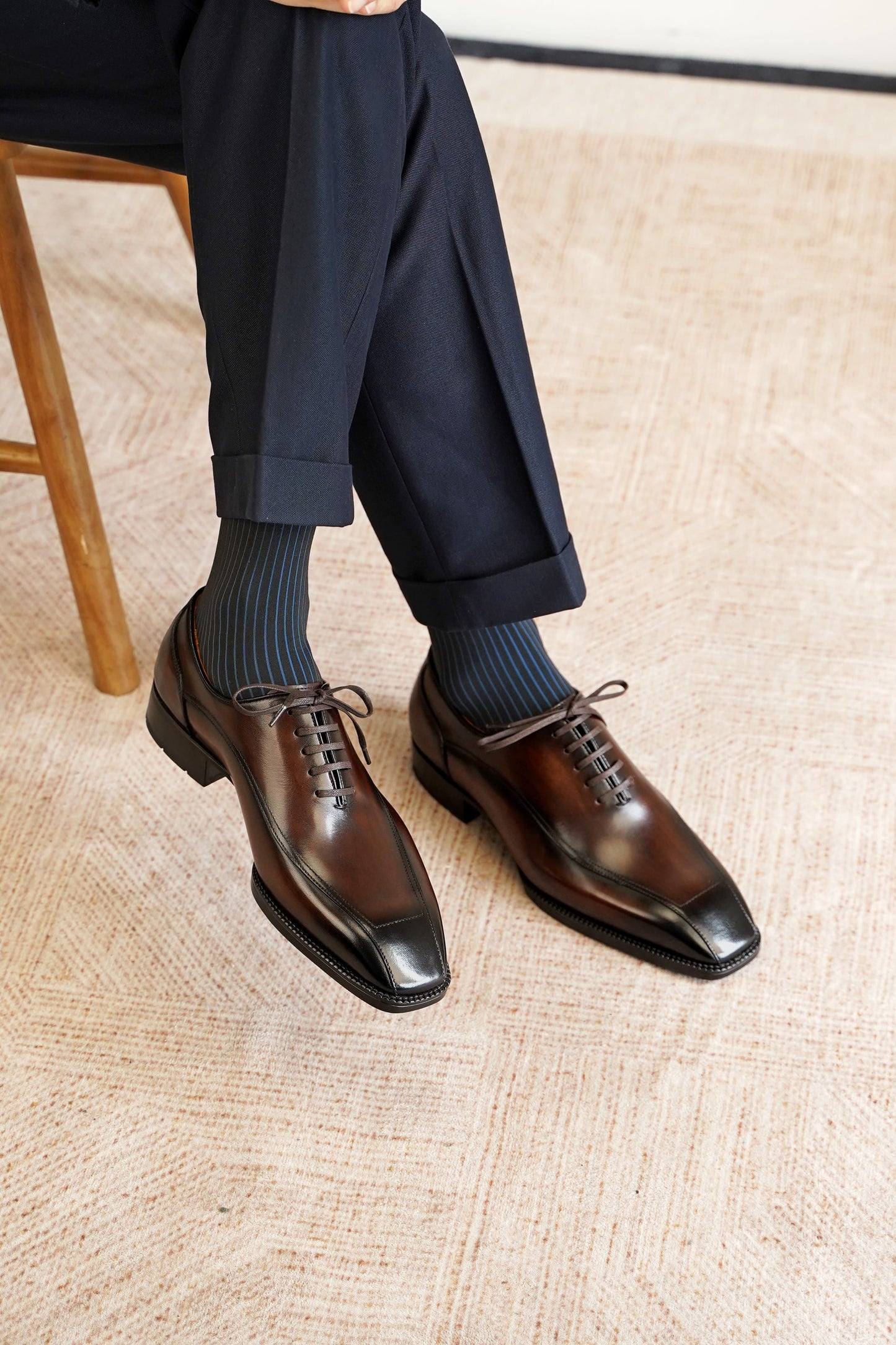 A close-up of a person's legs wearing dark navy trousers, dark blue ribbed socks, and polished dark brown oxford shoes with black toe caps, seated on a wooden stool over a textured beige rug.