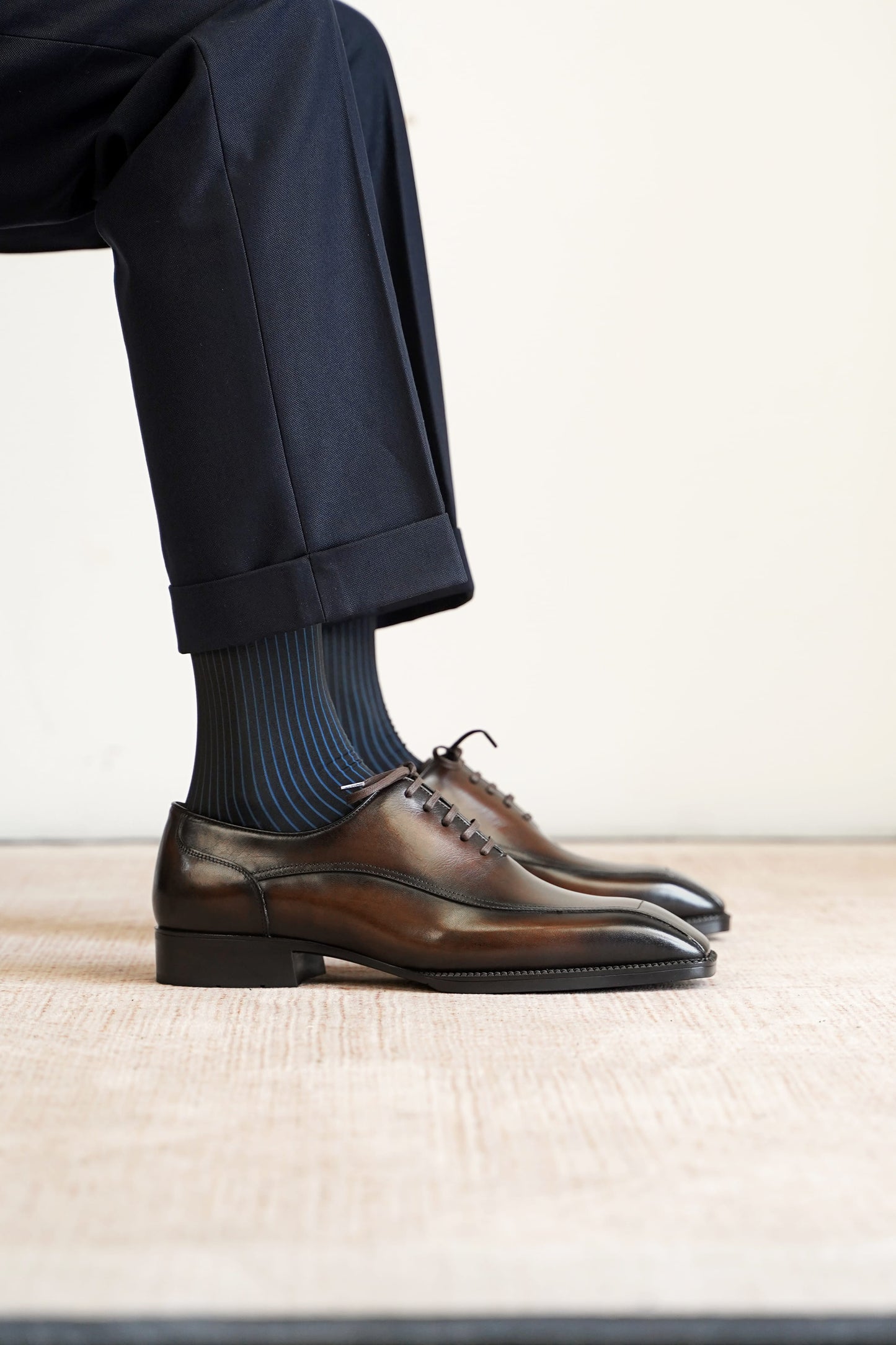 A close-up of a person's legs wearing dark navy trousers, blue striped socks, and dark brown leather oxford dress shoes on a light beige textured floor against a plain off-white wall.