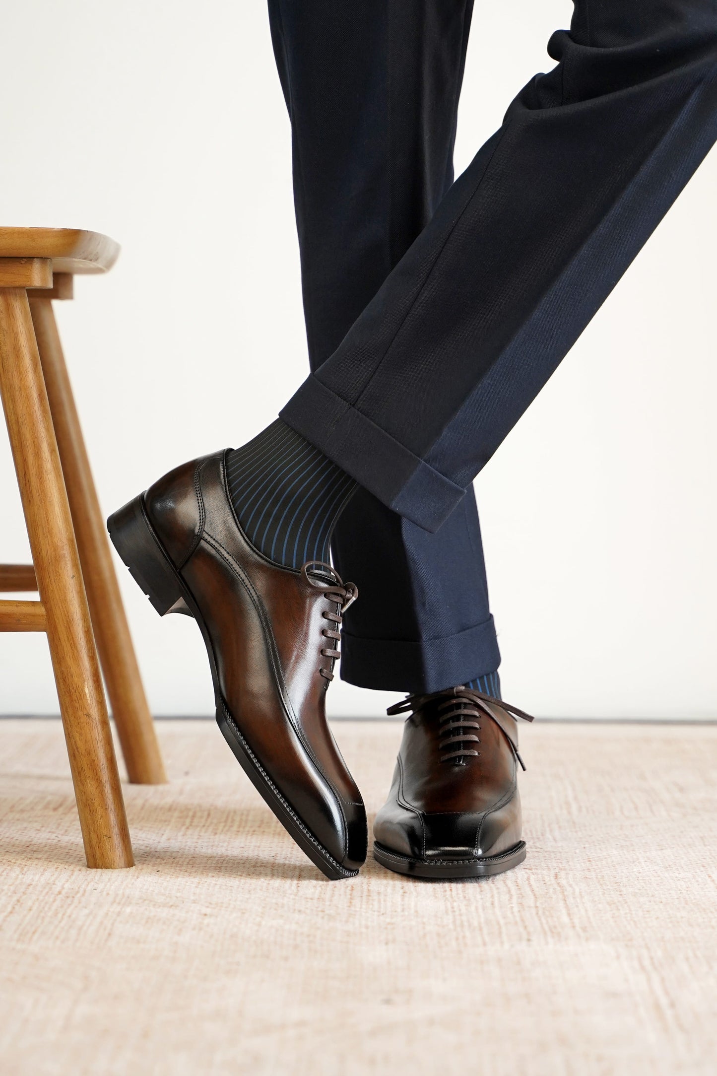 A close-up of a person's legs wearing dark brown oxford dress shoes with dark gray striped socks and navy trousers, standing on a light beige textured floor next to a wooden stool.