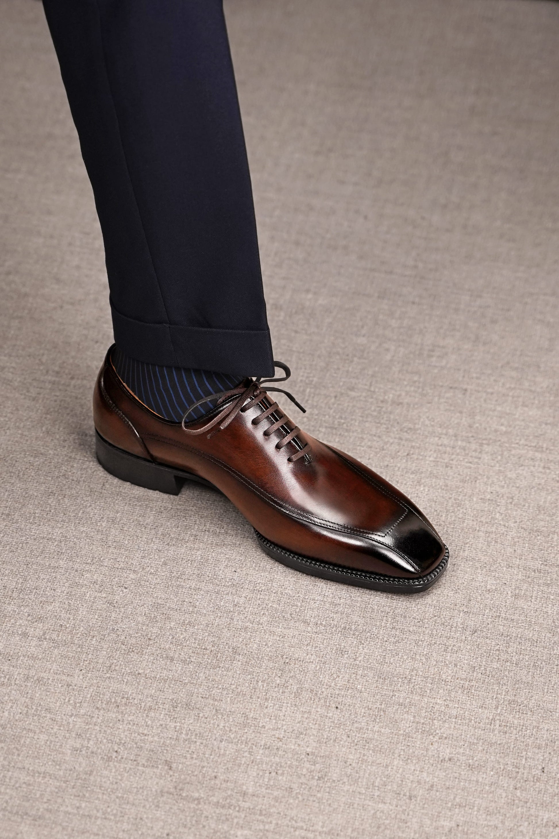 A close-up of a man's foot wearing a polished brown oxford dress shoe with a glossy black toe cap, paired with navy trousers and striped socks, standing on a textured beige carpet.