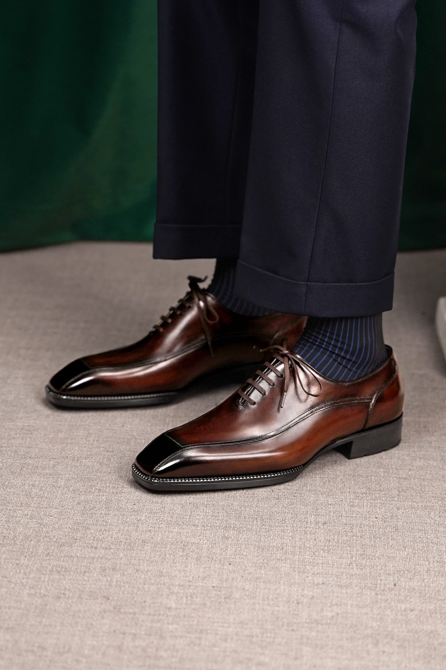 A pair of men's brown leather wingtip oxford shoes with a glossy black toe cap, worn with dark blue socks and navy trousers on a textured beige floor.