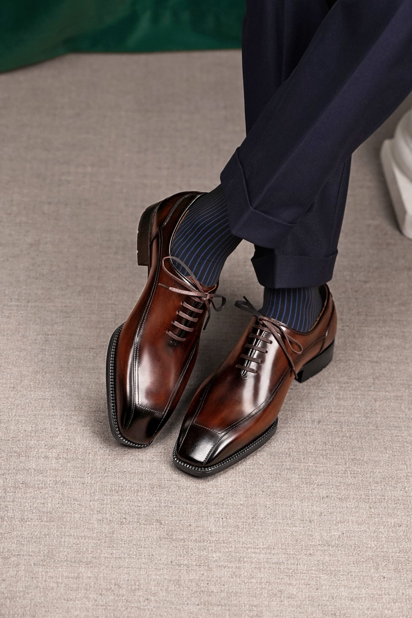A pair of men's brown leather oxford dress shoes with a glossy cap toe, worn with dark navy trousers and dark blue striped socks on a textured beige carpet.