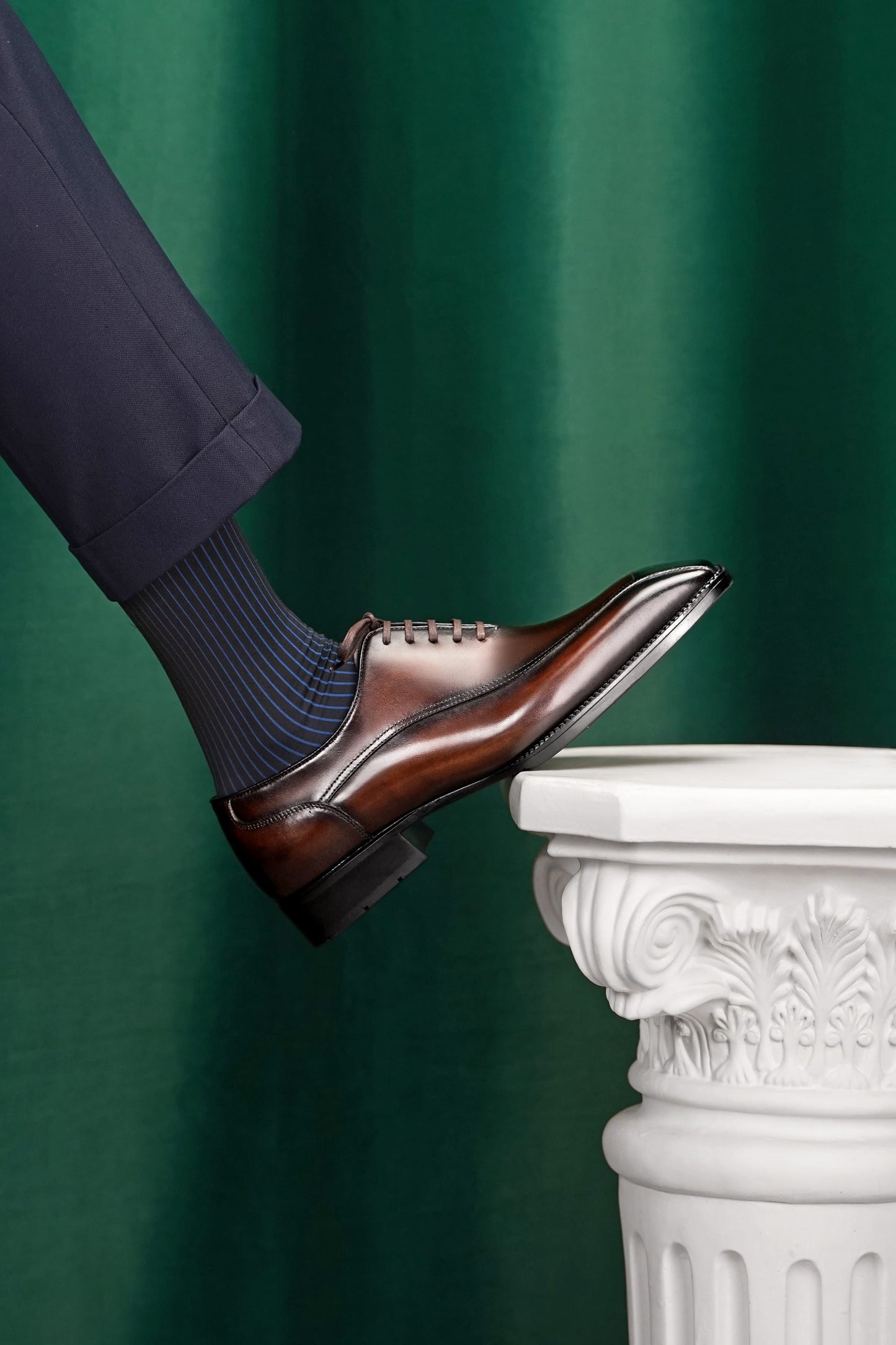 A man's foot wearing a brown leather oxford dress shoe with dark blue ribbed socks, stepping onto a white decorative column against a dark green curtain backdrop.