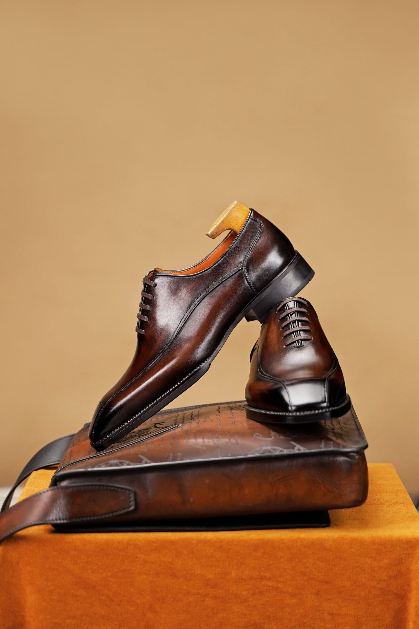A pair of dark brown leather lace-up dress shoes displayed on top of a matching brown leather bag against a warm tan studio background.